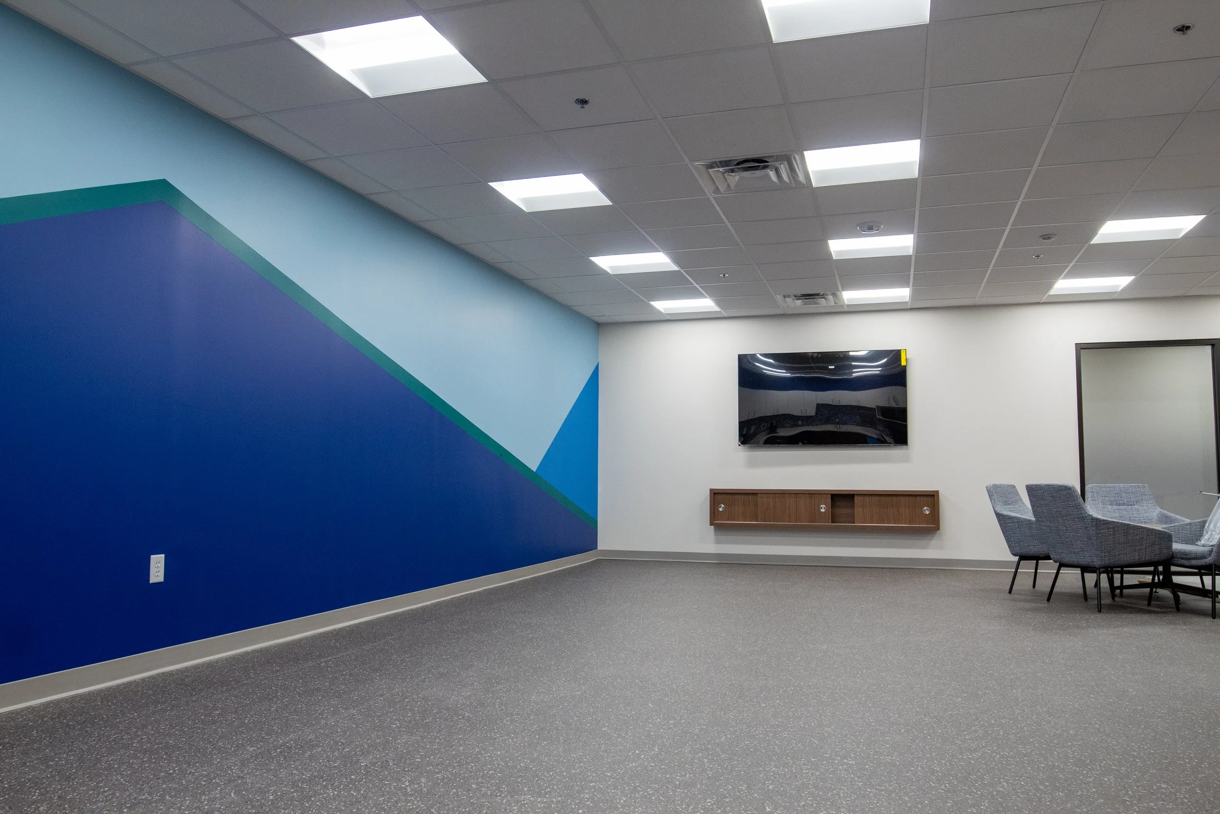 Empty modern conference room with blue and white walls, gray carpet, ceiling lights, wall-mounted TV, and gray chairs around a table.