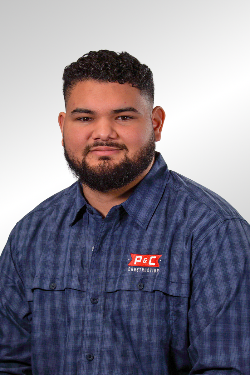 Portrait of a young man with dark curly hair and a beard, wearing a blue plaid shirt with a P&C Construction logo, against a plain white background.