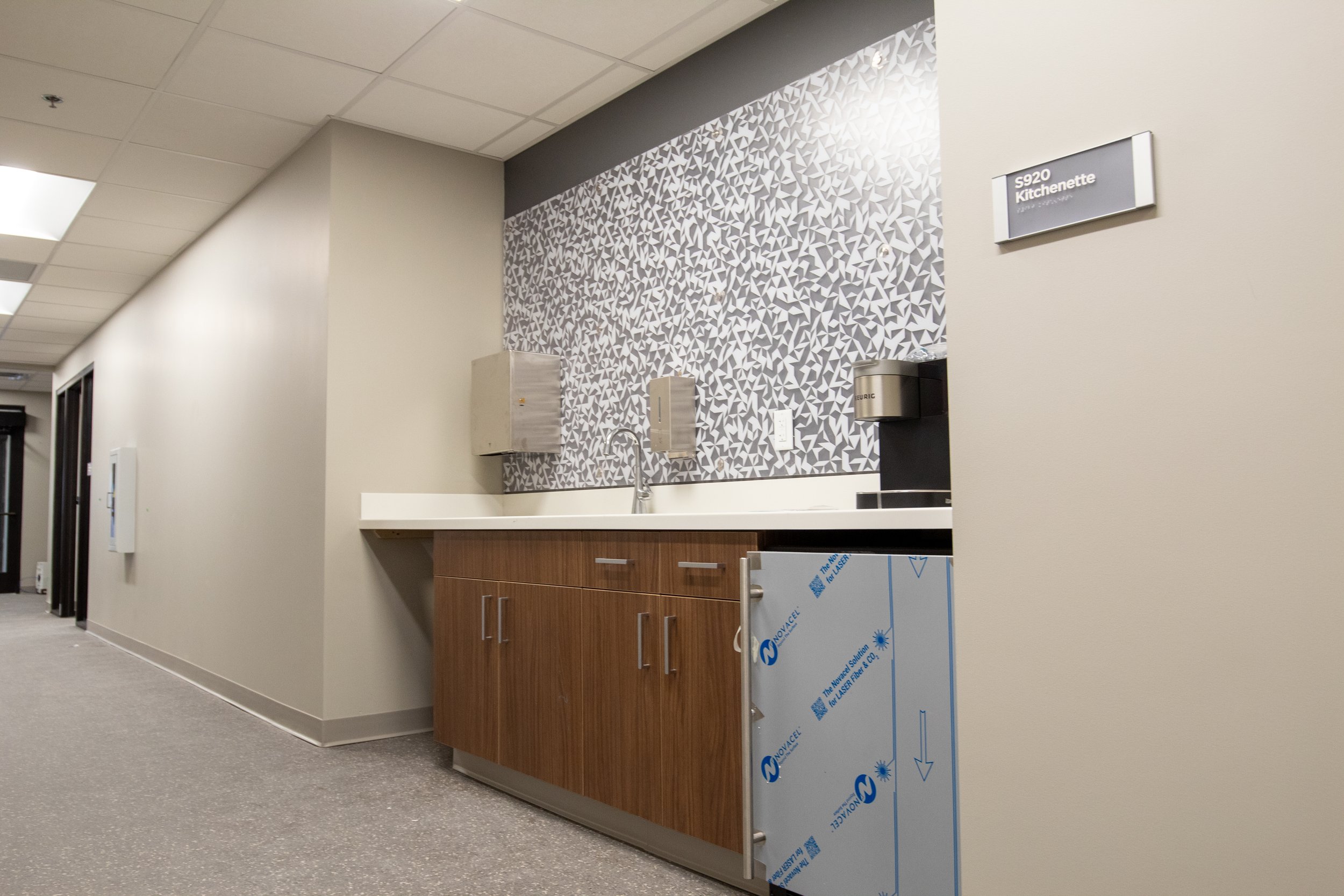 Empty kitchenette area with a patterned wall, wooden cabinets, a sink, a paper towel dispenser, and a coffee machine in a building corridor.