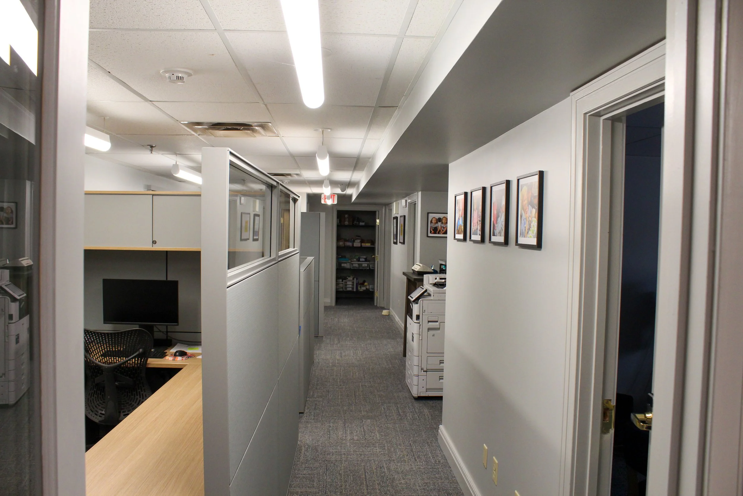 Office hallway with cubicles, framed photographs on the wall, a photocopier, and a door leading to another room.