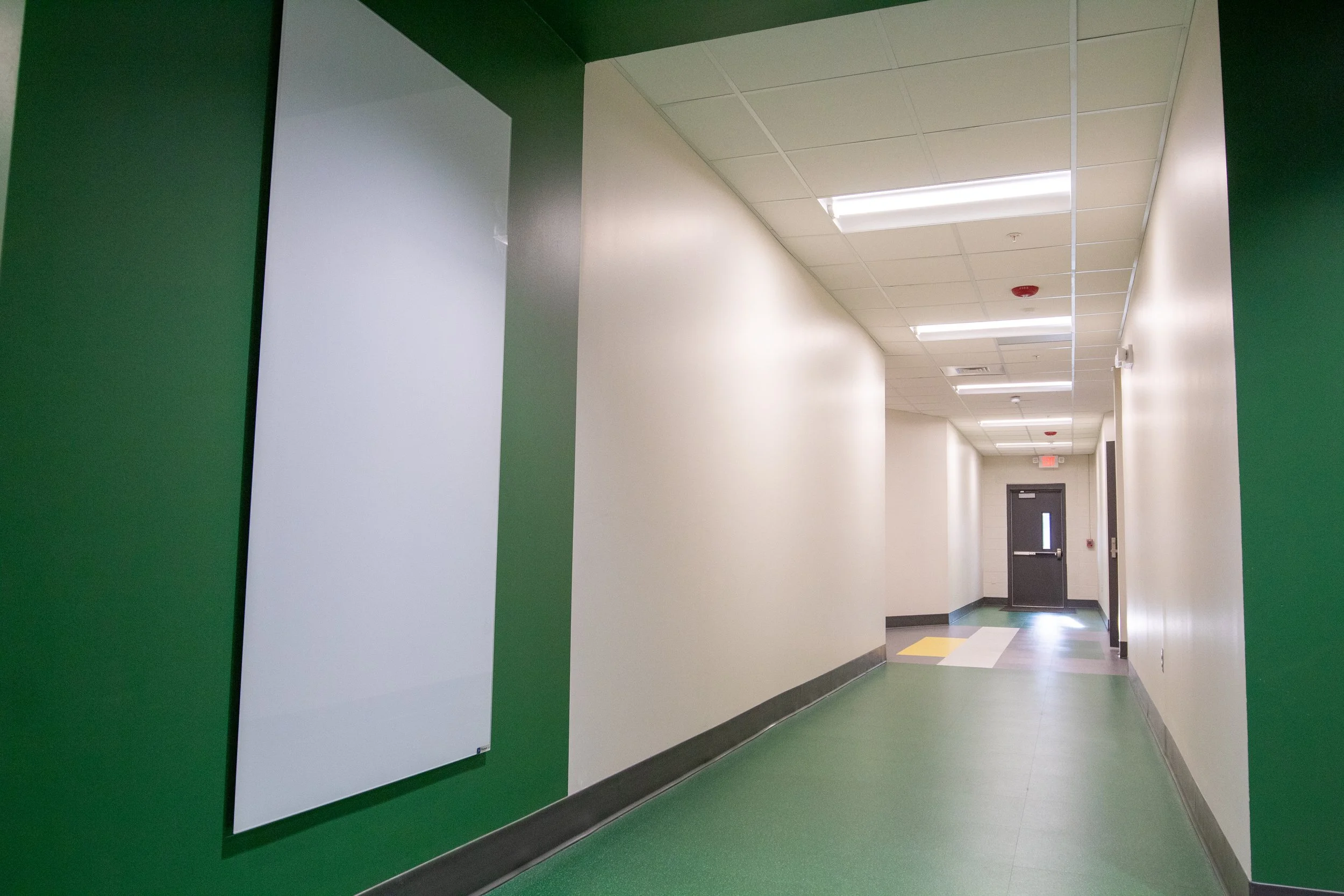 Empty hospital hallway with white and green walls, fluorescent lighting, and a door at the end.