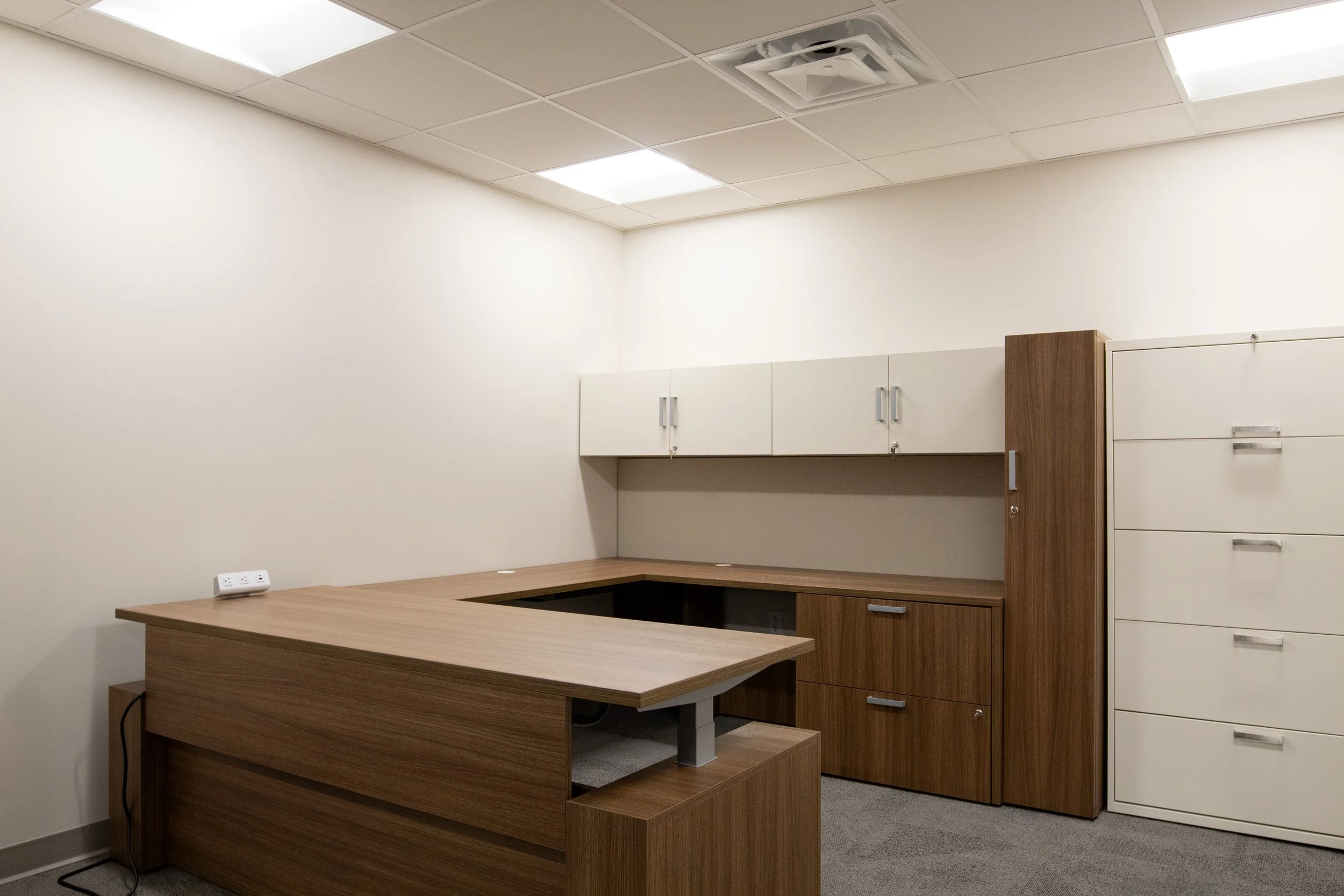 Empty office with wooden L-shaped desk, white cabinets on wall, and a filing cabinet in the corner.
