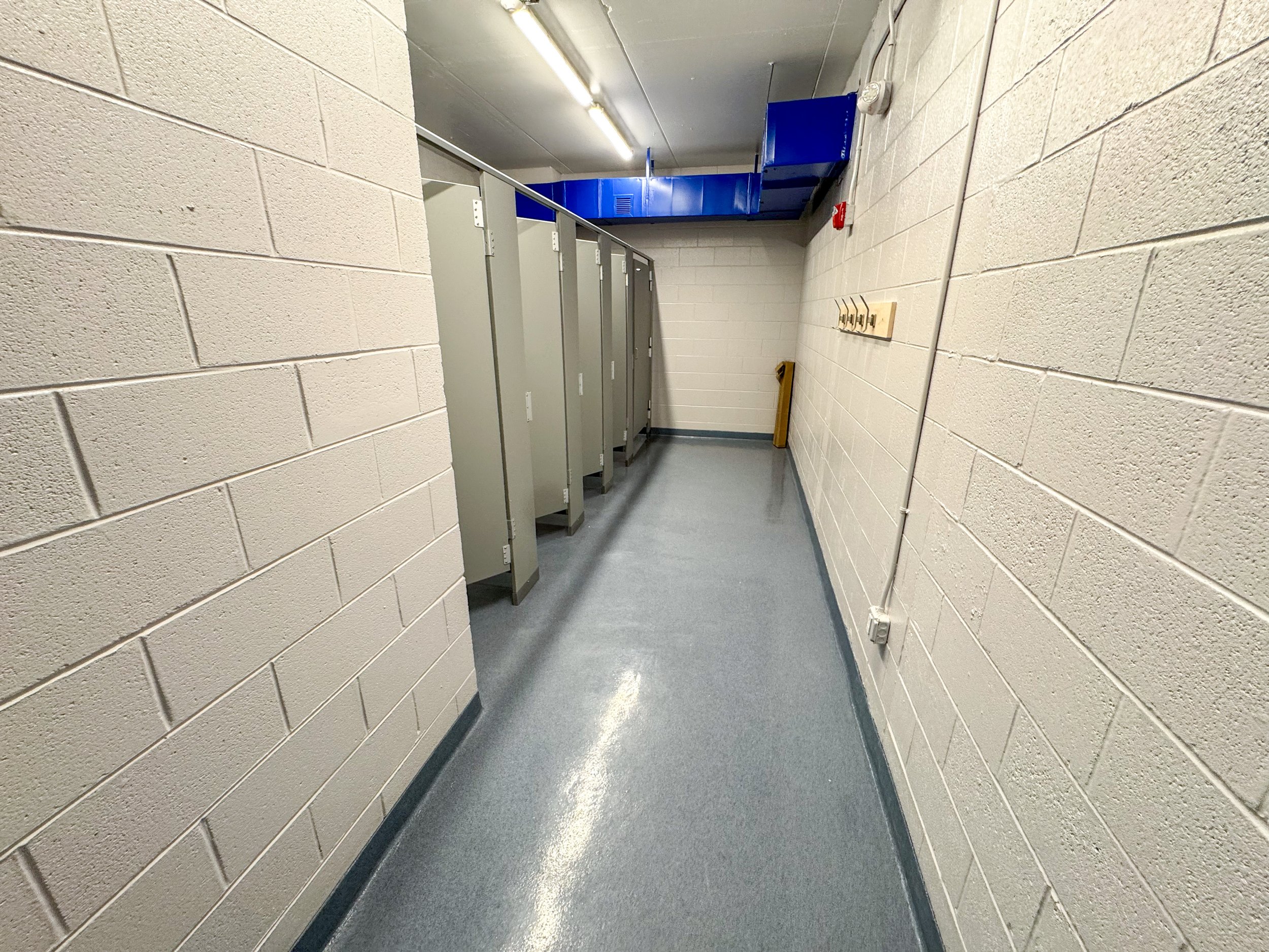 Public restroom with beige cinder block walls, grey flooring, several light grey toilet stalls, a row of hooks on the right wall, and blue ductwork on the ceiling.
