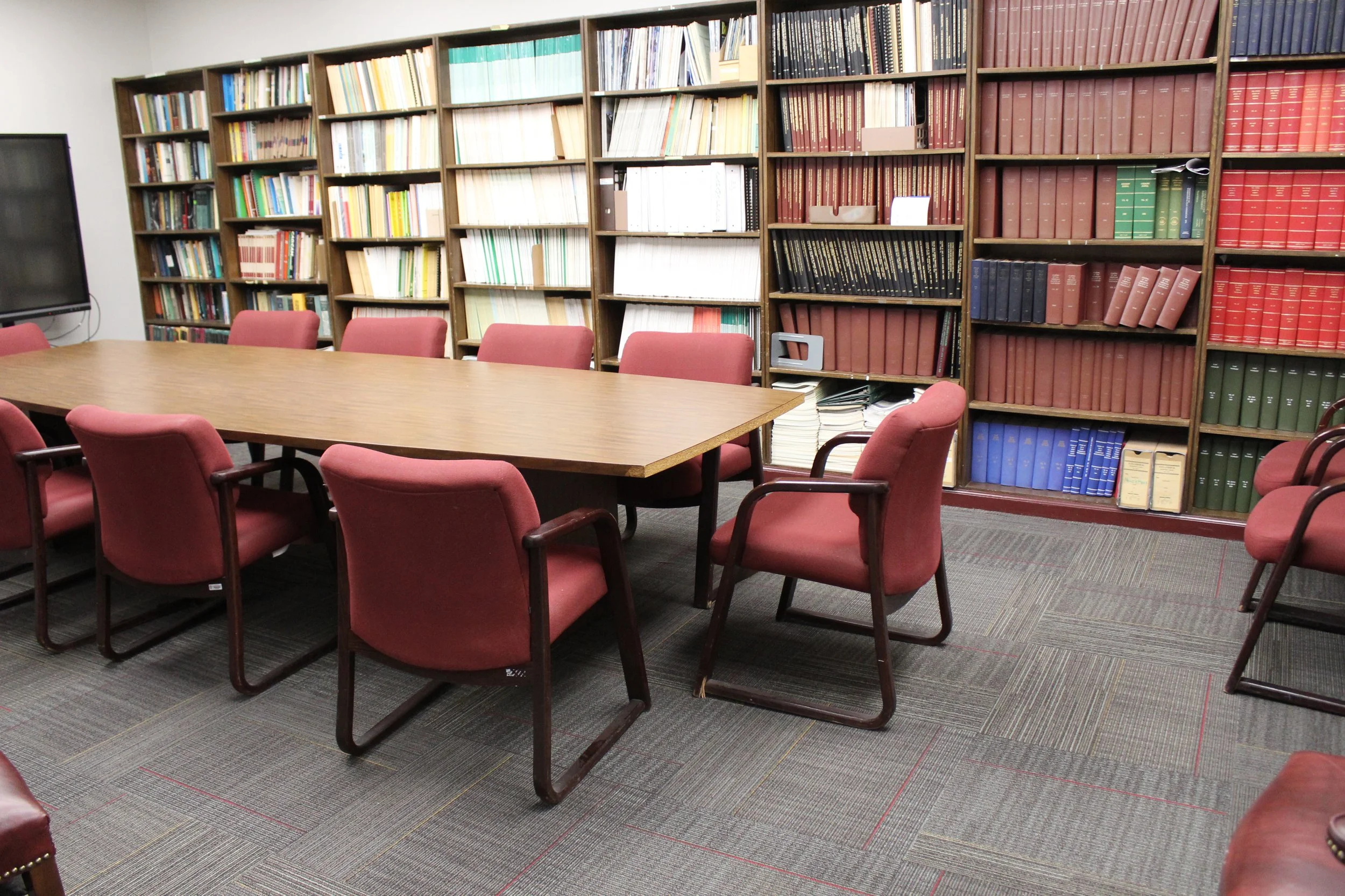 A conference room with a long wooden table surrounded by red cushioned chairs, and filled bookshelves with various books and files against the wall.