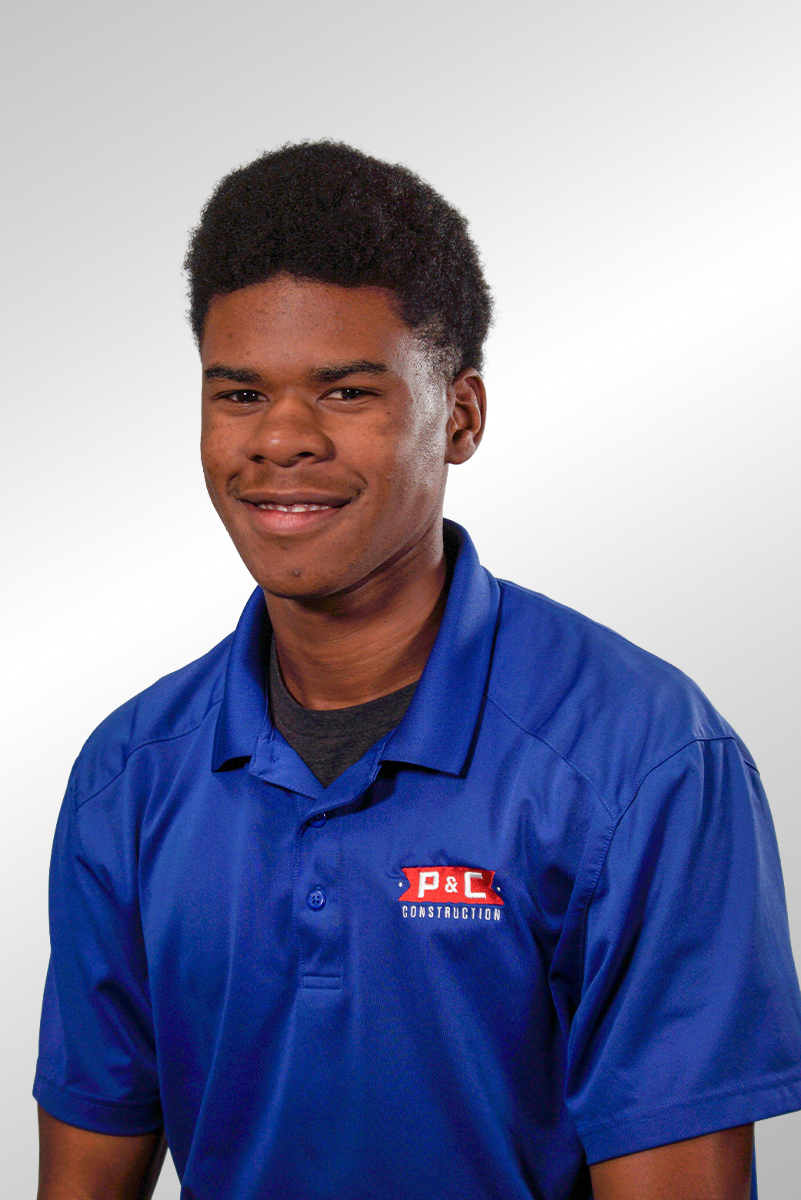 A young man with short black hair and dark skin, wearing a blue polo shirt with a P&C Construction logo, smiling at the camera against a plain white background.