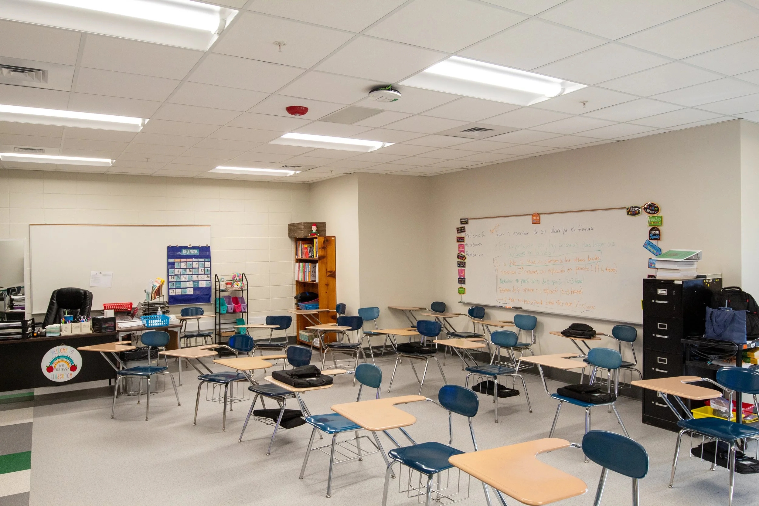 Empty classroom with desks and chairs, whiteboard, bookshelves, and teaching supplies.