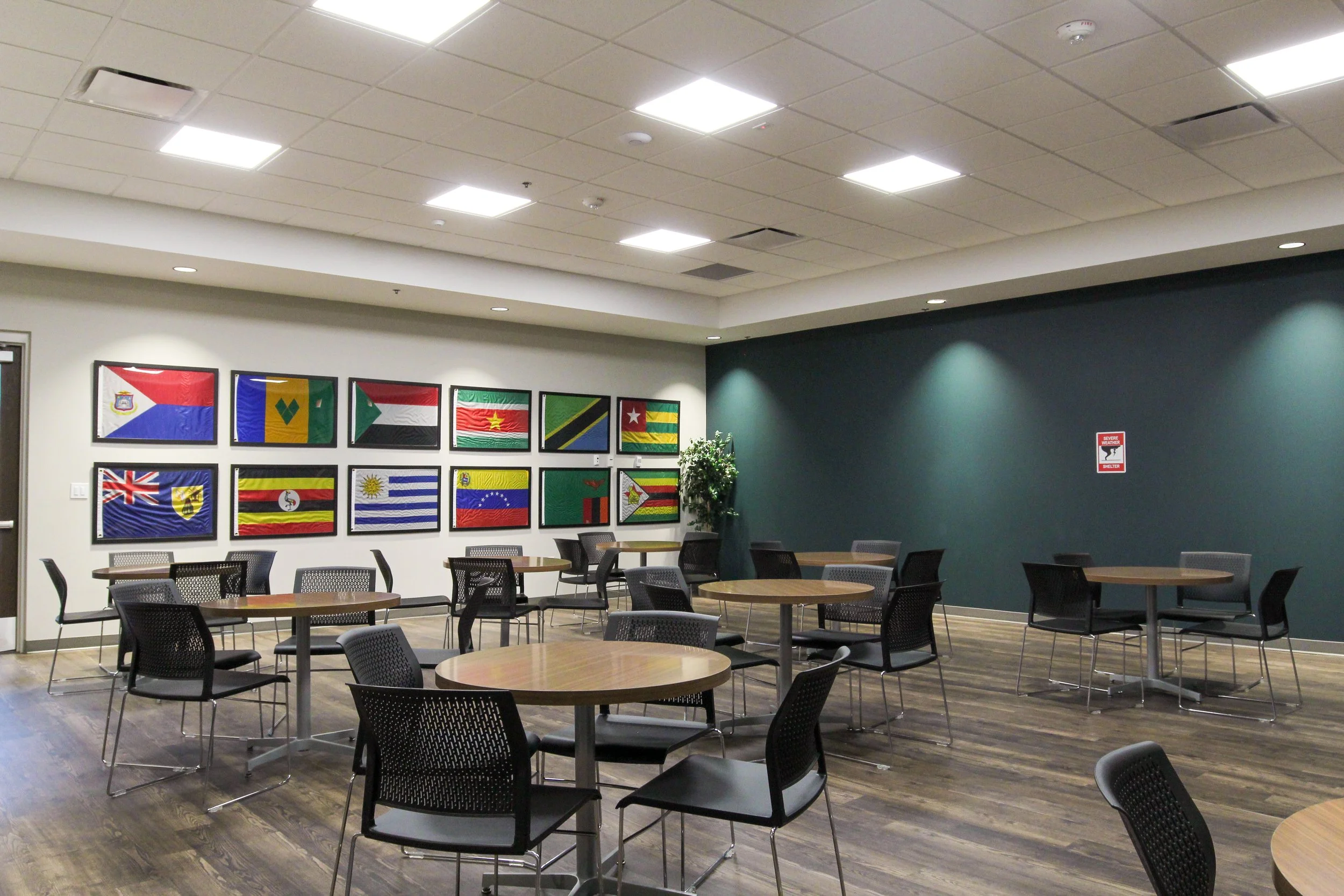 Indoor meeting or dining area with round tables and black chairs, wood floors, walls adorned with framed flags from various countries, and a notice about severe weather shelter on the wall. ORCM design build completed by  P&C Construction, Inc.