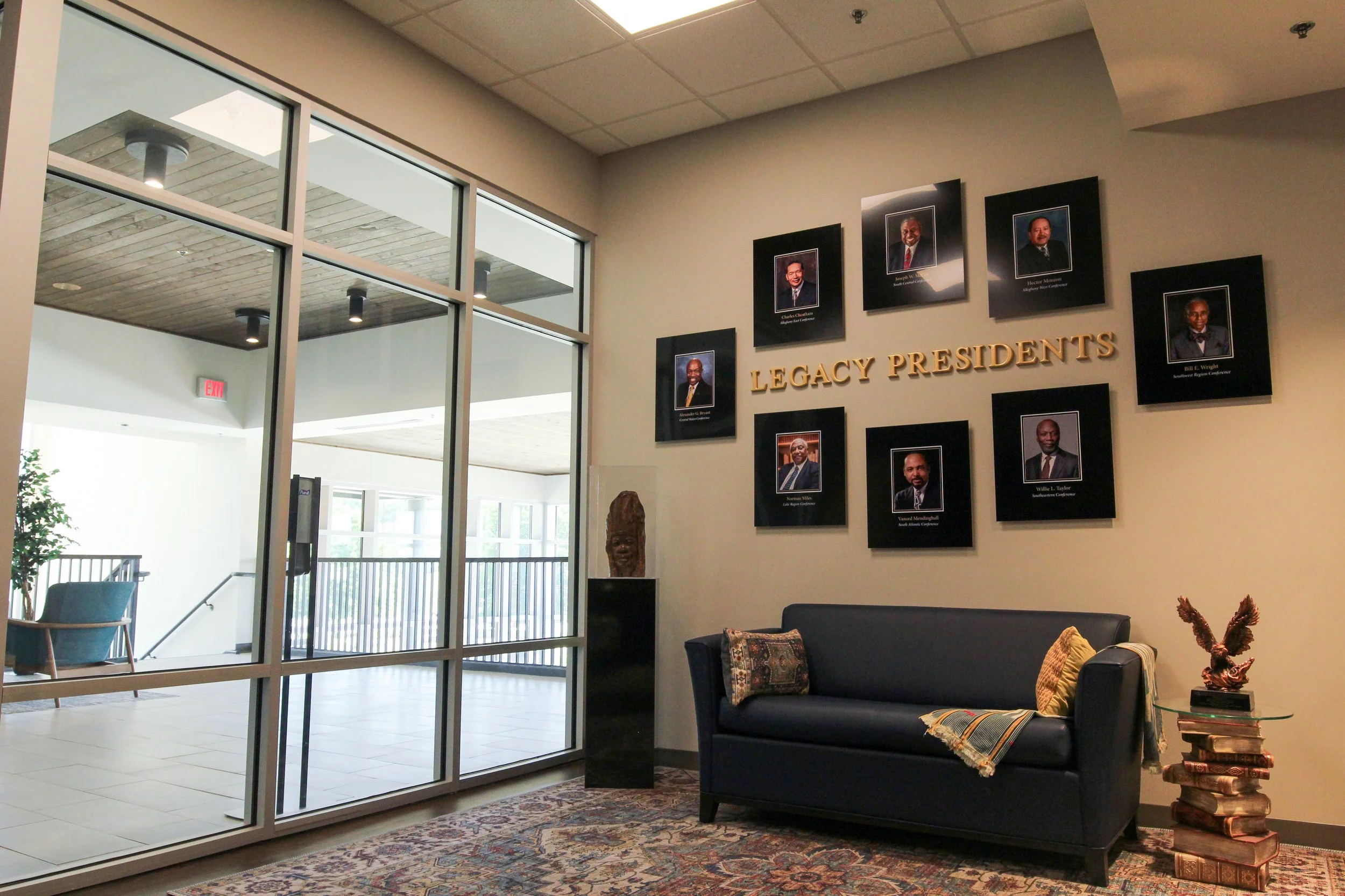 Lobby area with glass doors, framed portraits on the wall labeled 'Legacy Presidents', black sofa with decorative pillows, a small side table with a stack of books and a brass sculpture, against a beige wall and carpeted floor. 