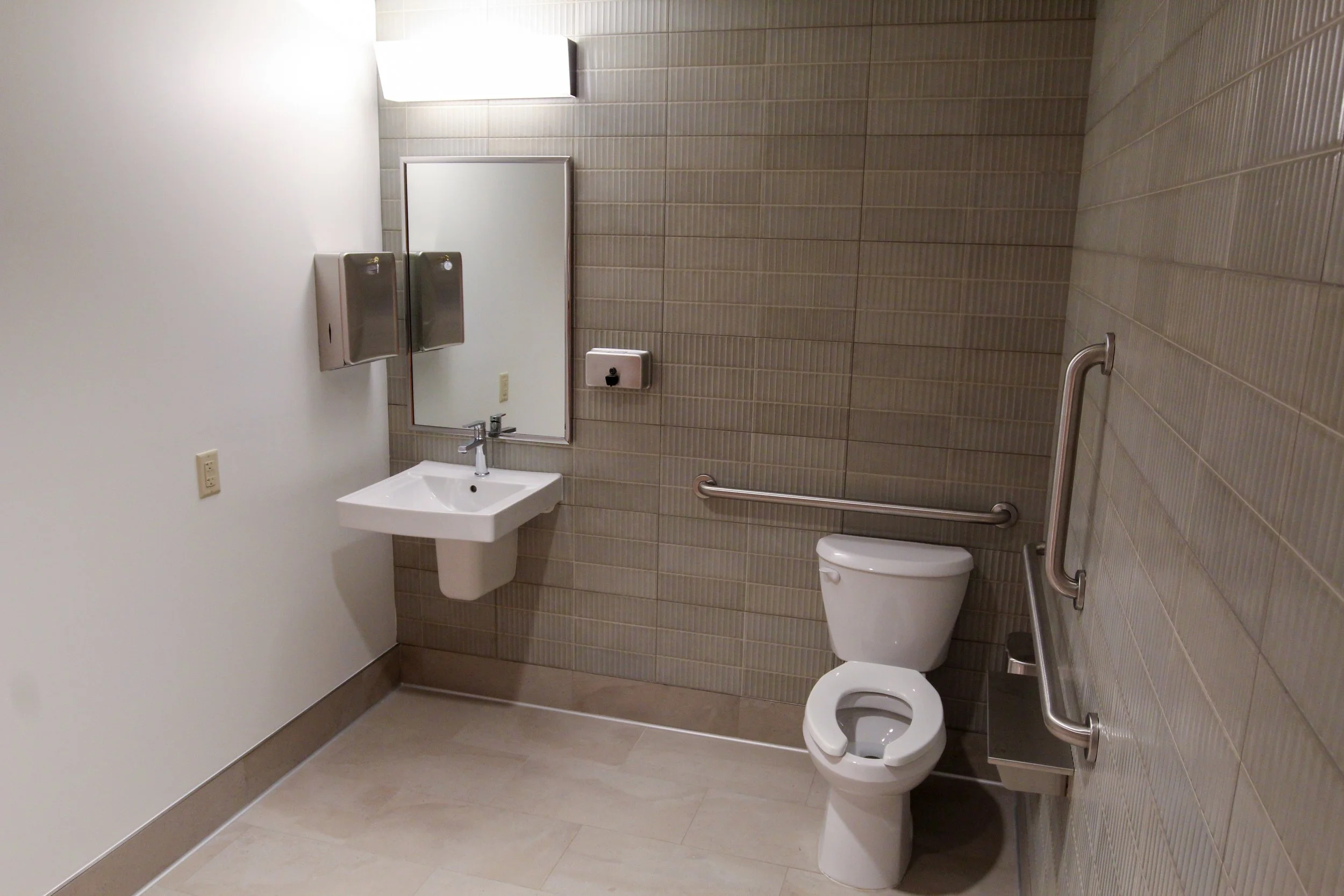 Accessible men's restroom with beige tiled walls, a white toilet, a small white sink, a mirror, hand dryers, a mirror, and guardrails for support.