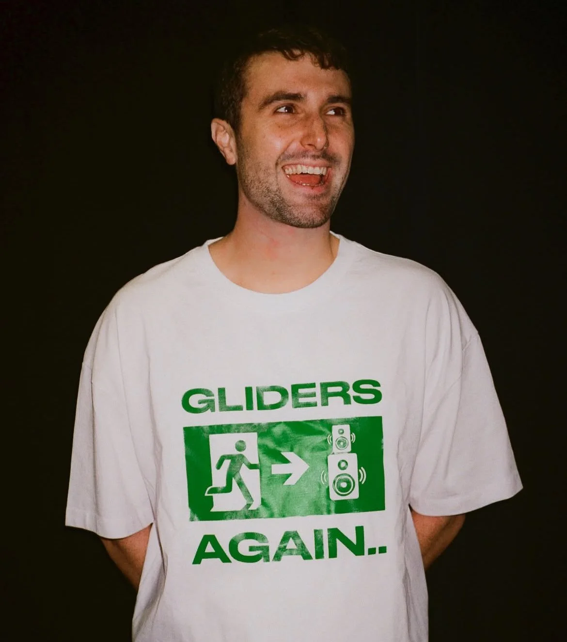 A man wearing a white T-shirt with green graphics and text that says 'GLIDERS AGAIN.' The shirt features a person running towards a speaker with sound waves.