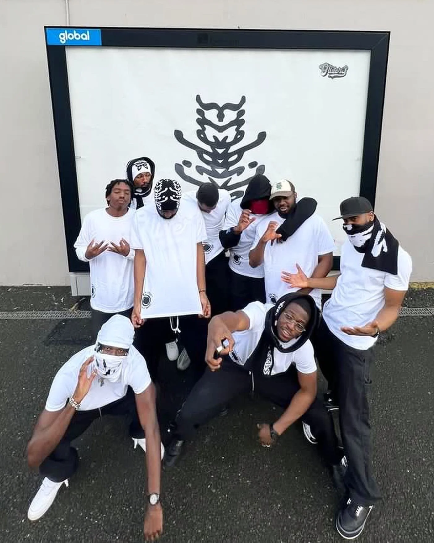 Group of young men wearing white and black clothing, some with bandanas and masks, posing in front of a large tiger face graphic on a display board outdoor. They are striking various poses, with some kneeling and others standing.