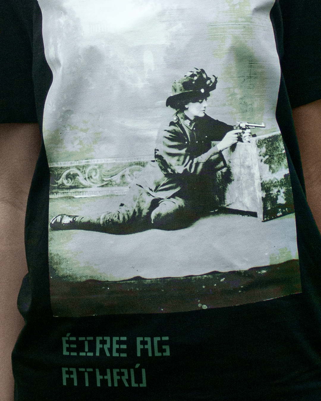 A person wearing a black t-shirt with a printed image of a historical woman in a 19th-century dress and hat kneeling and aiming a pistol, with the words 'ÉTRE AG ATHRÚ' at the bottom.