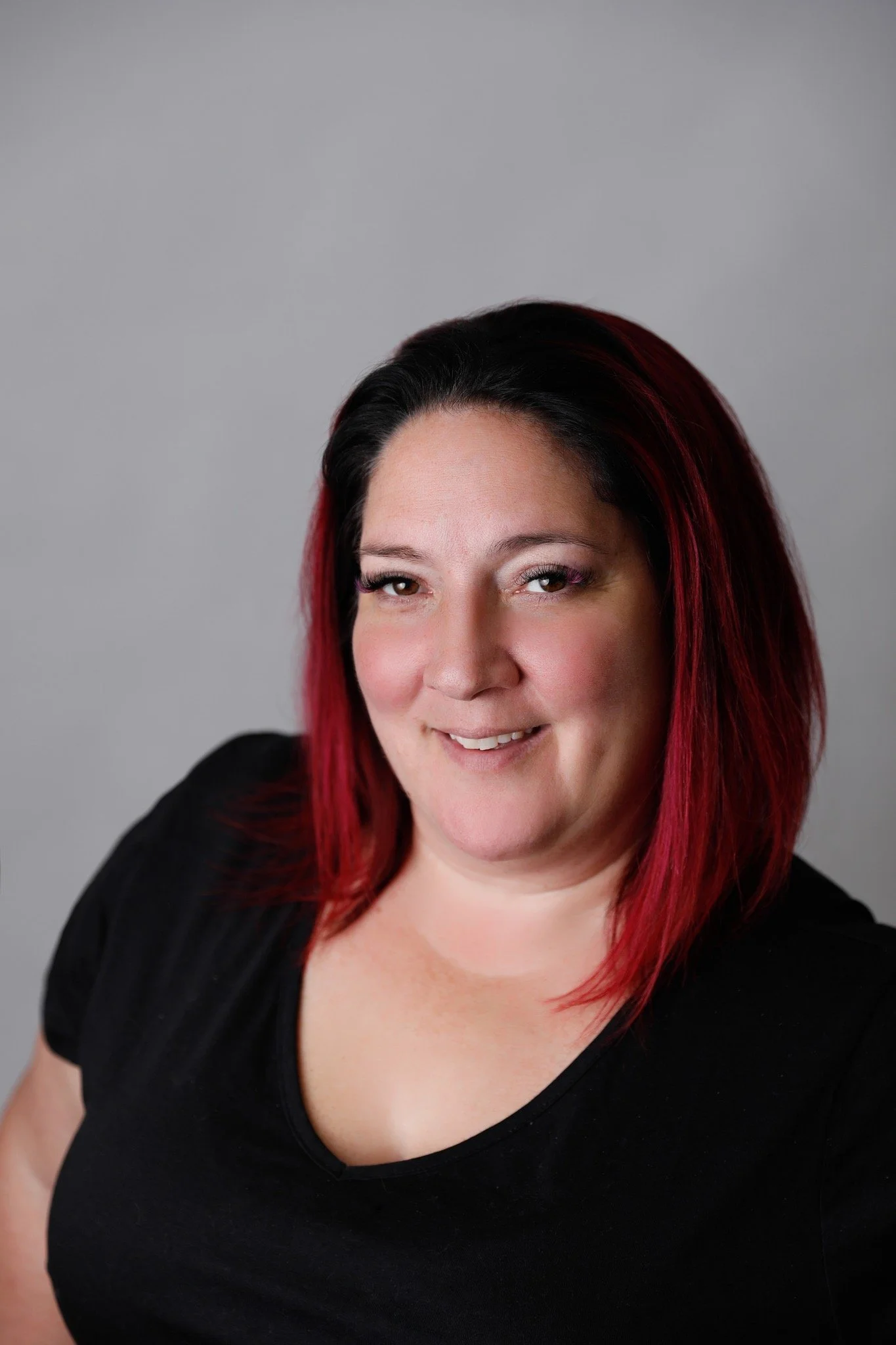 A woman with shoulder-length red and black hair, smiling, wearing a black top, against a plain gray background.