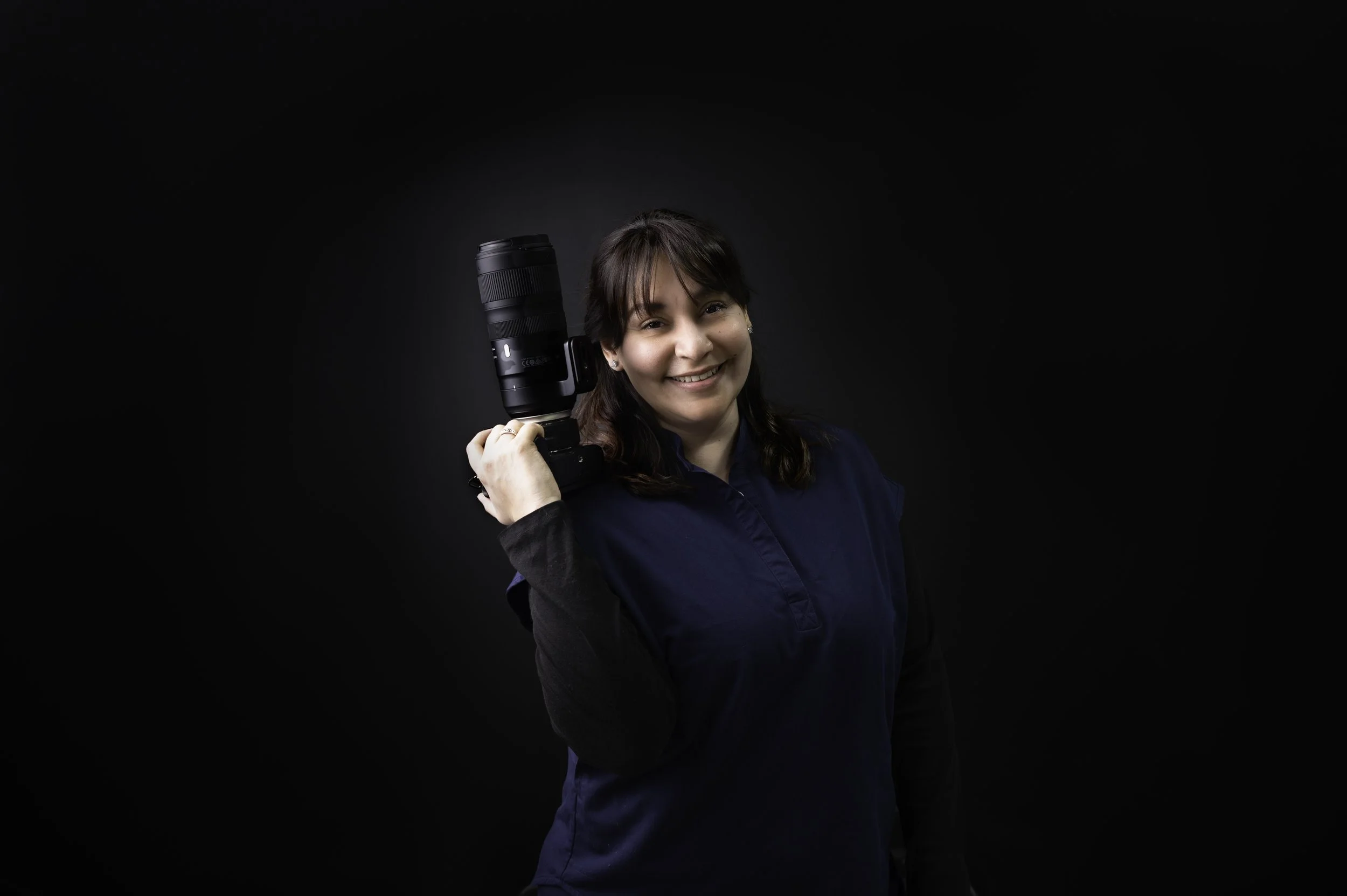 Woman holding a professional camera with a large lens on her shoulder, smiling, against a dark background.