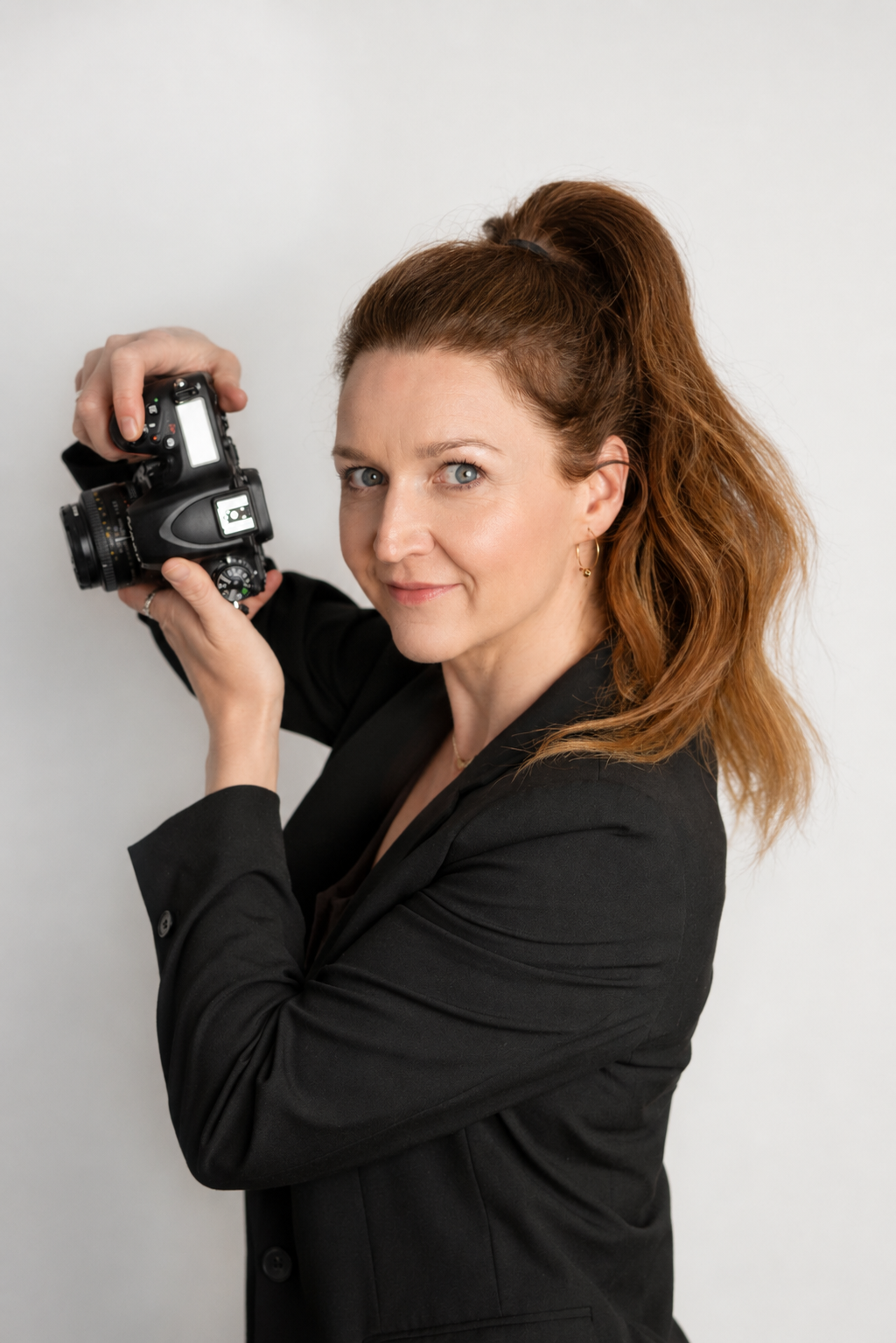 A woman with long brown hair holding an old-fashioned camera, standing against a dark background.