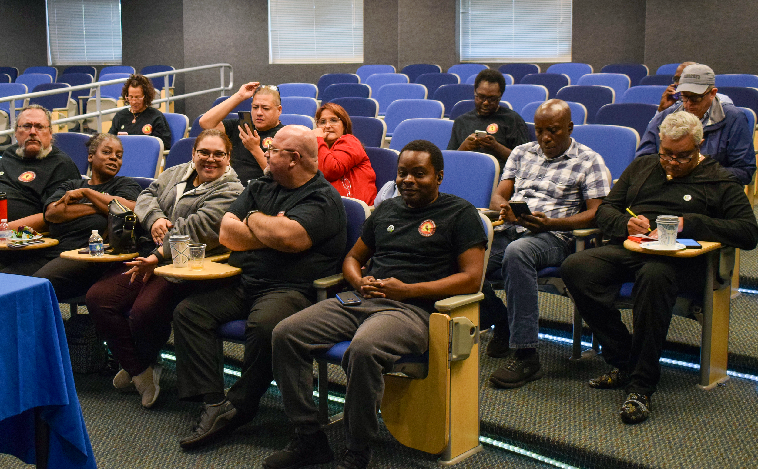 Group of people sitting in an auditorium, some using their phones, some taking notes, with blue chairs and windows in the background.