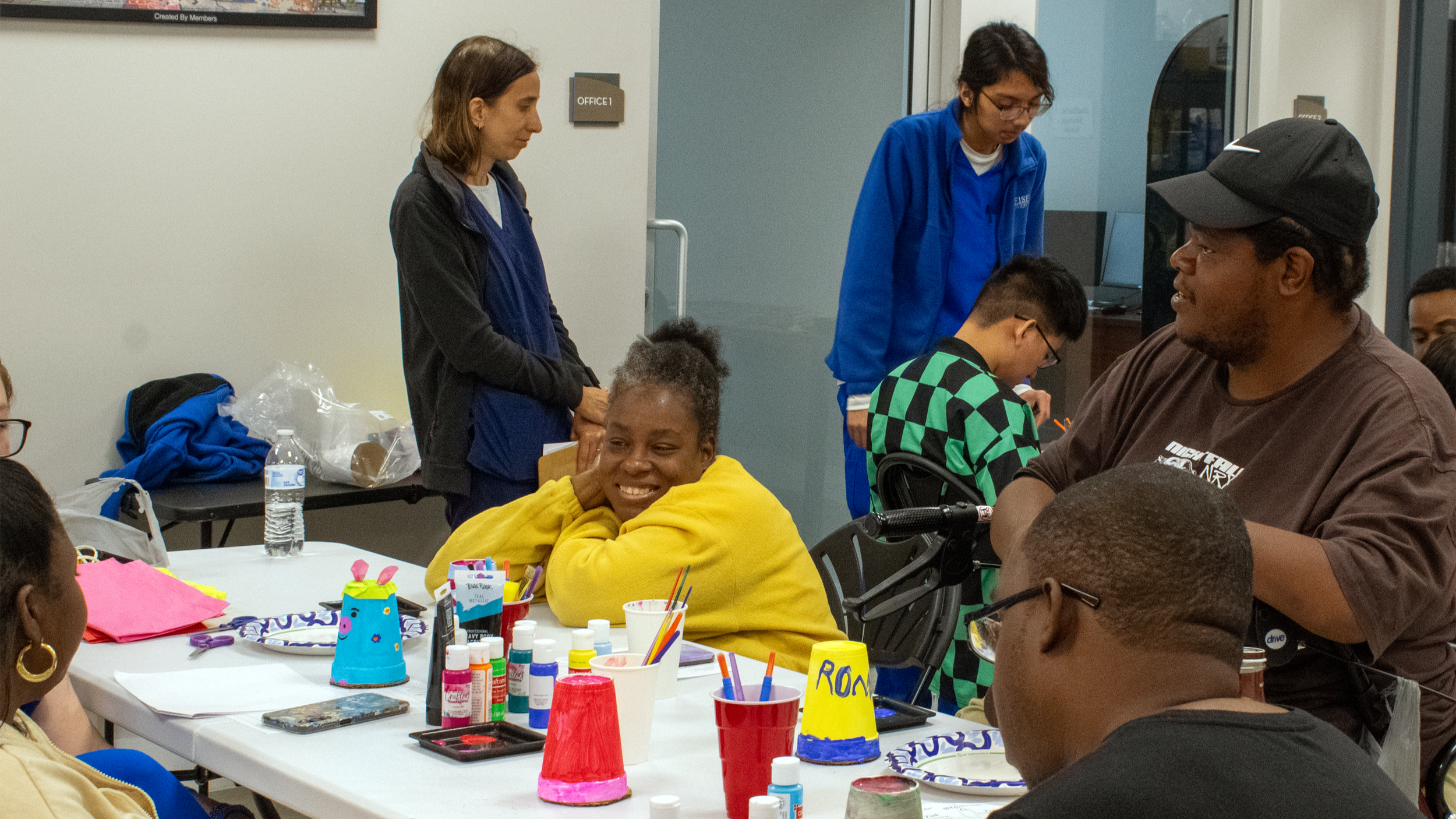 Group of people sitting around a table with arts and crafts supplies, including paint bottles, brushes, and paper; some people are smiling and engaging in conversation.