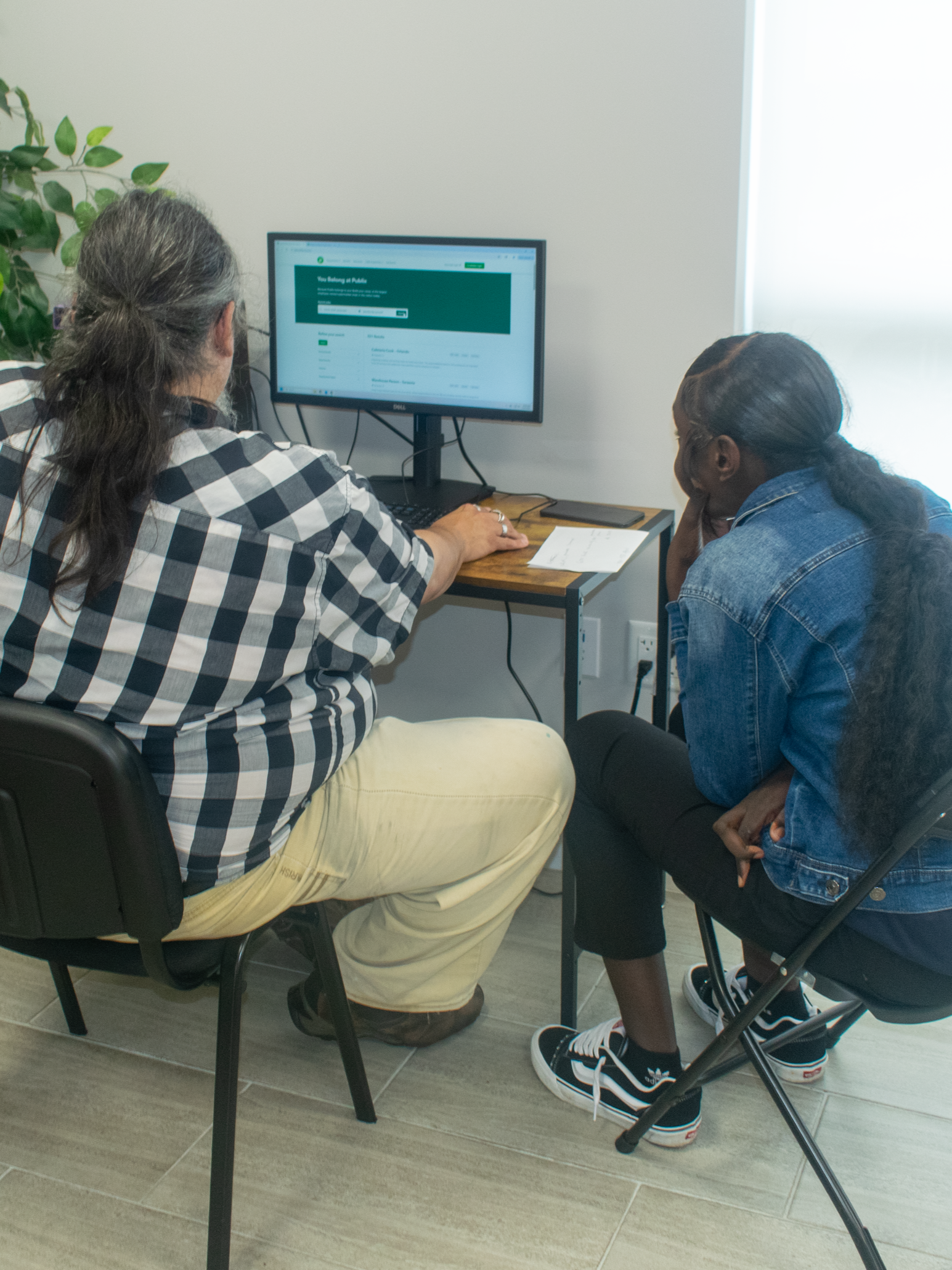 Two women sitting at a small desk, looking at a computer monitor, in an office setting.