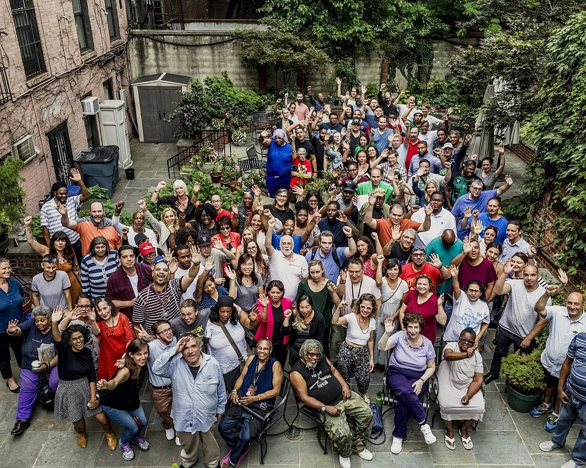 A large diverse group of people gathered in an outdoor courtyard, posing for a photo with many raising their hands or waving. The setting includes plants, outdoor furniture, and brick walls.