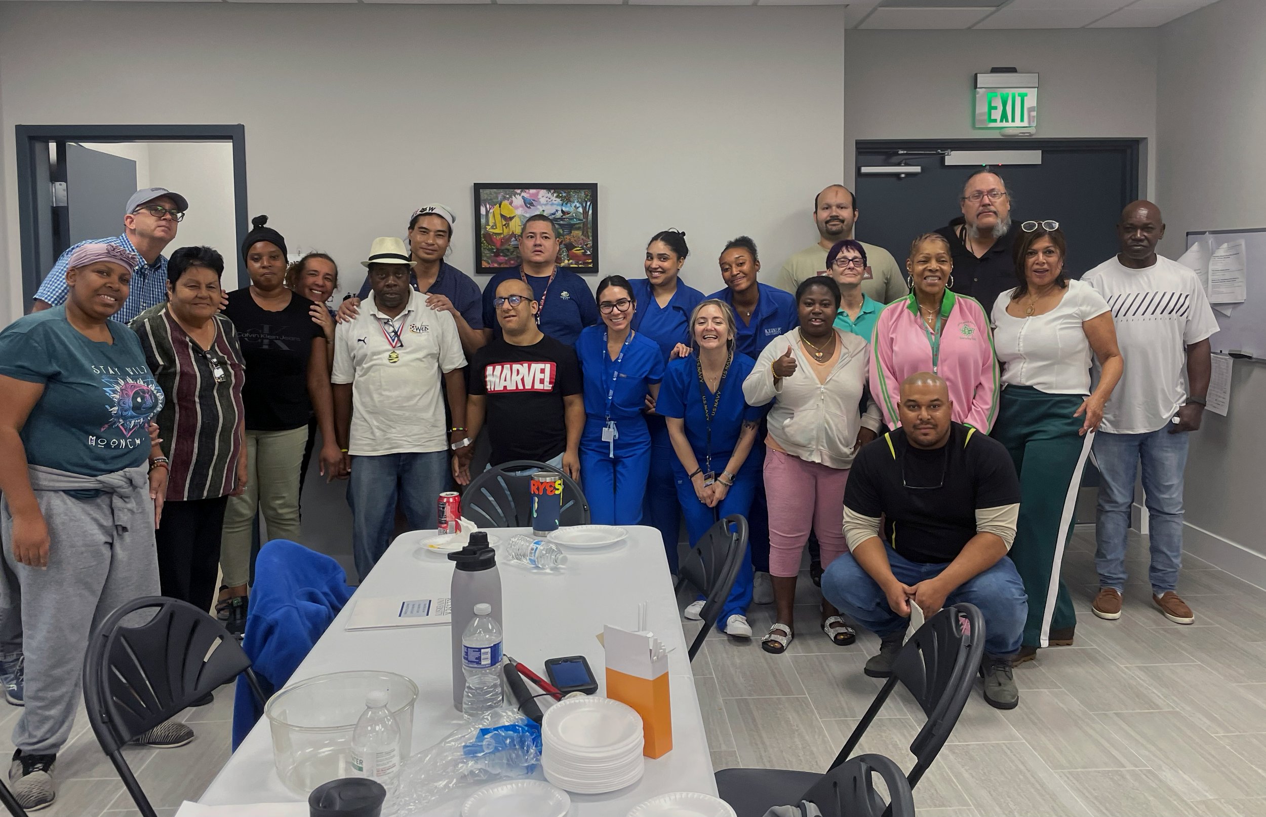 A diverse group of people posing indoors around a table, some wearing scrubs, indicating a medical or healthcare setting.