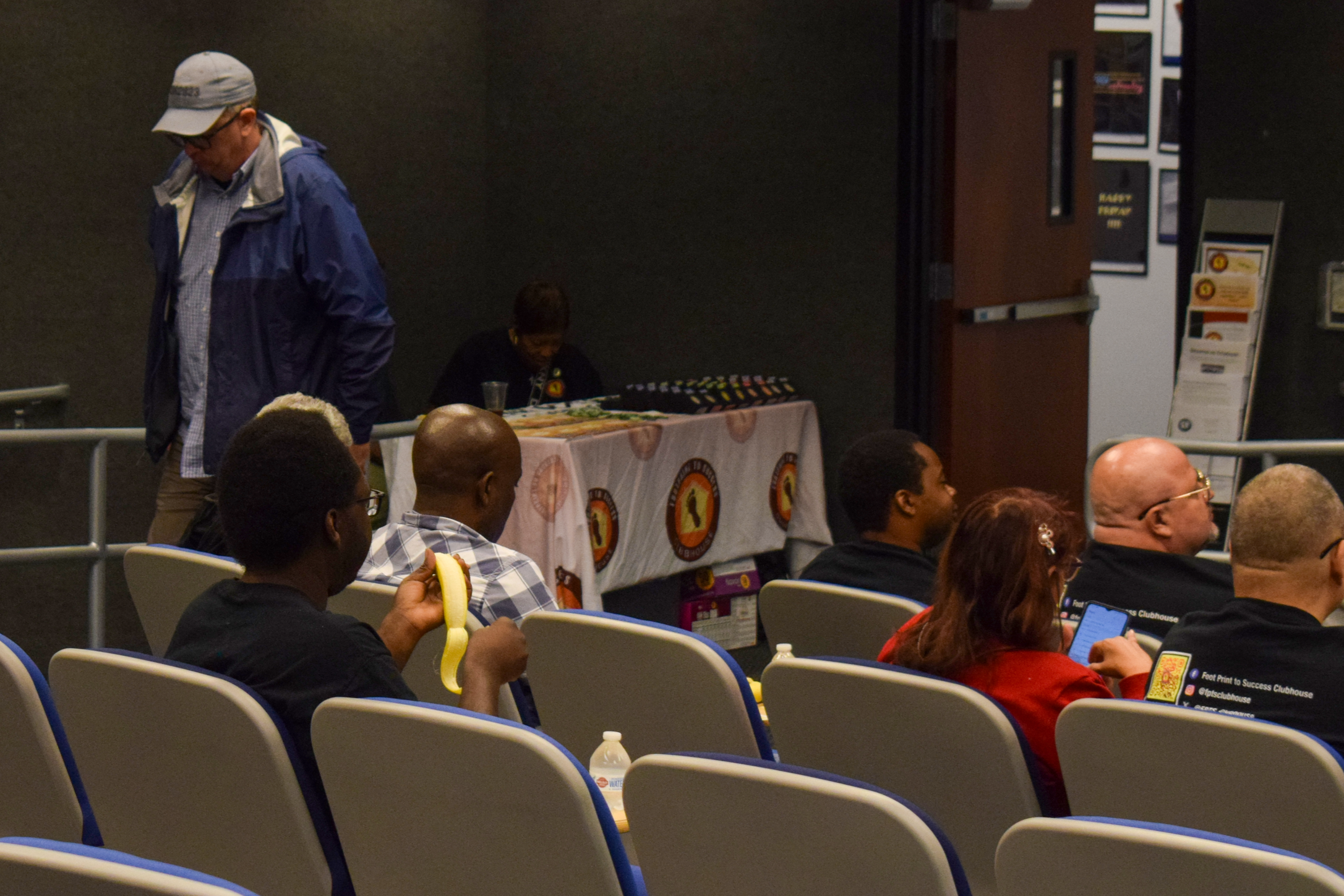 Audience seated in a conference room, some looking at their phones, a man holding a banana, a table with a cloth and a woman seated behind it, and an open door in the background.