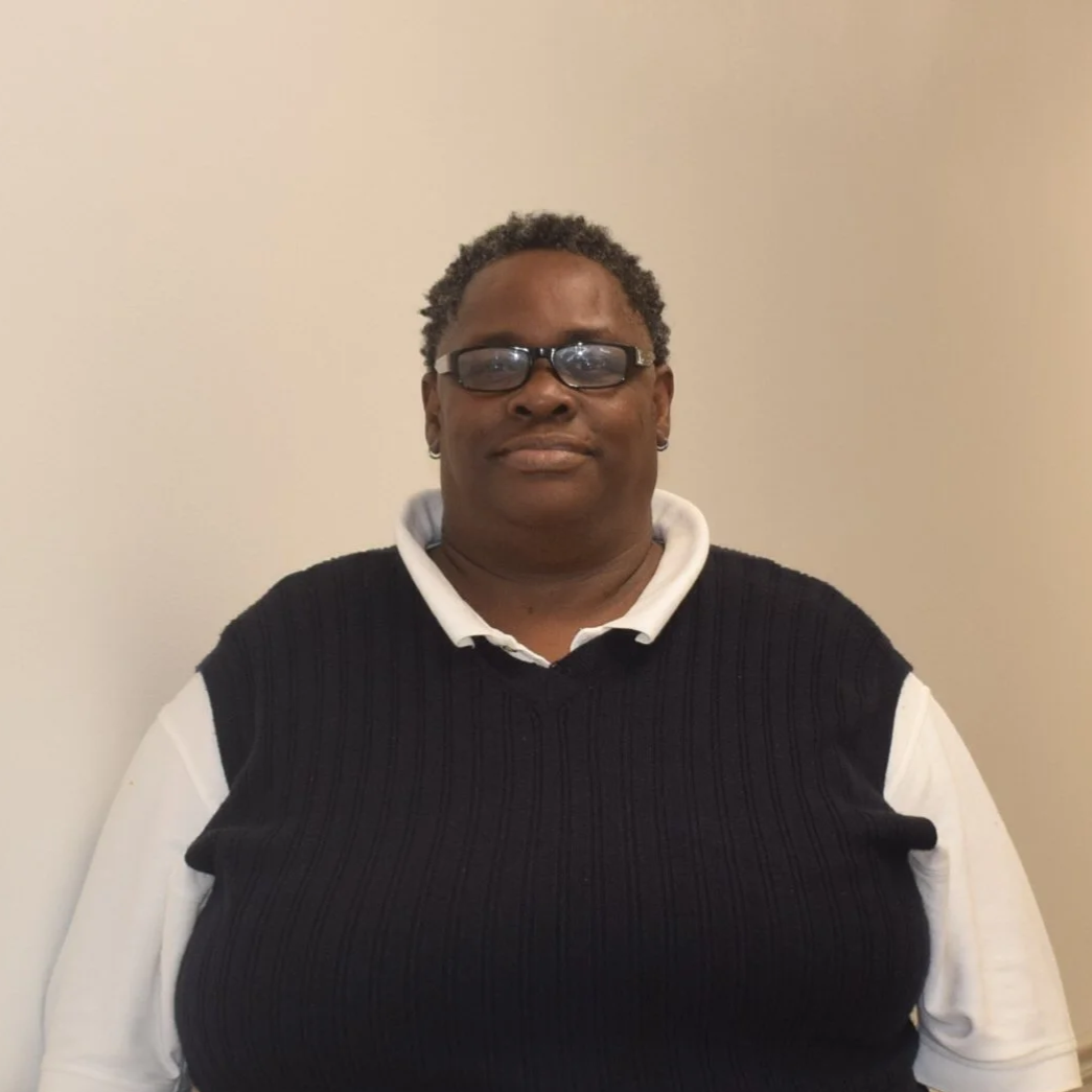 A woman with glasses and short curly hair, wearing a white collared shirt under a black sleeveless sweater, standing against a plain light-colored wall.