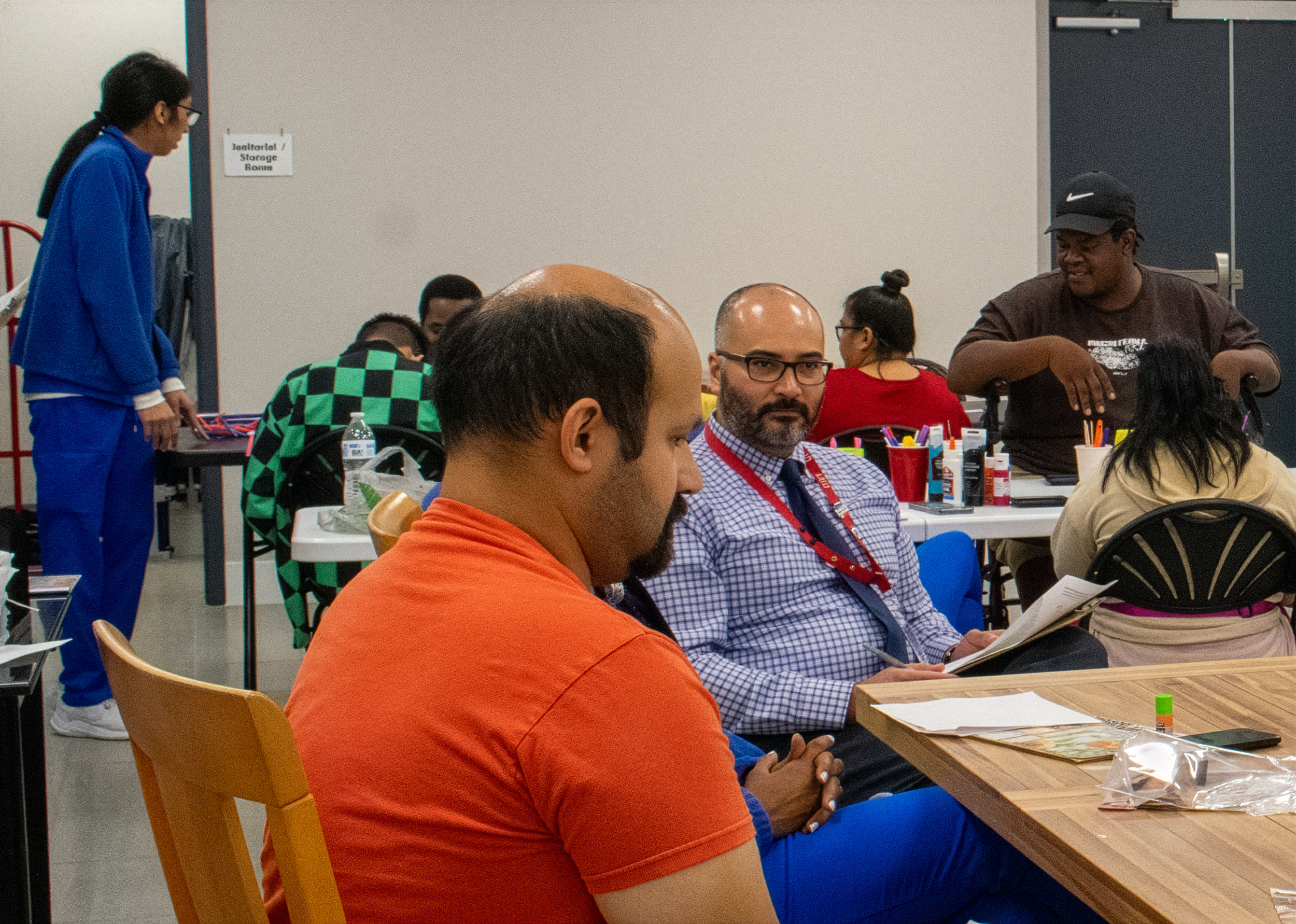 People sitting around a table in a classroom or workshop setting, engaging in discussion and activities. Some are taking notes, others are observing. Various art supplies and personal items are on the tables, and a sign on the wall reads 'Jacketotal Storage Room.'