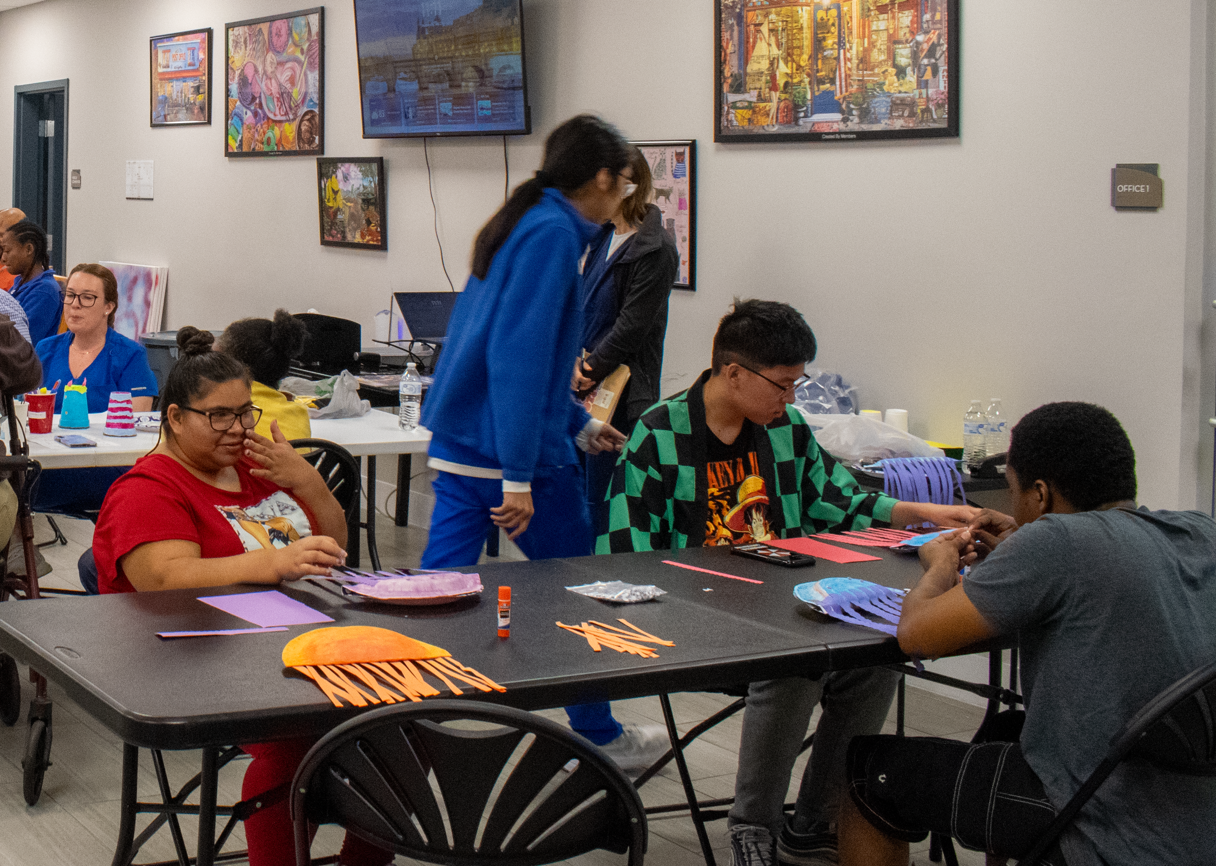 People participating in an arts and crafts workshop in a conference room, with colorful paper strips on the table, and artwork, computers, and water bottles visible in the background.