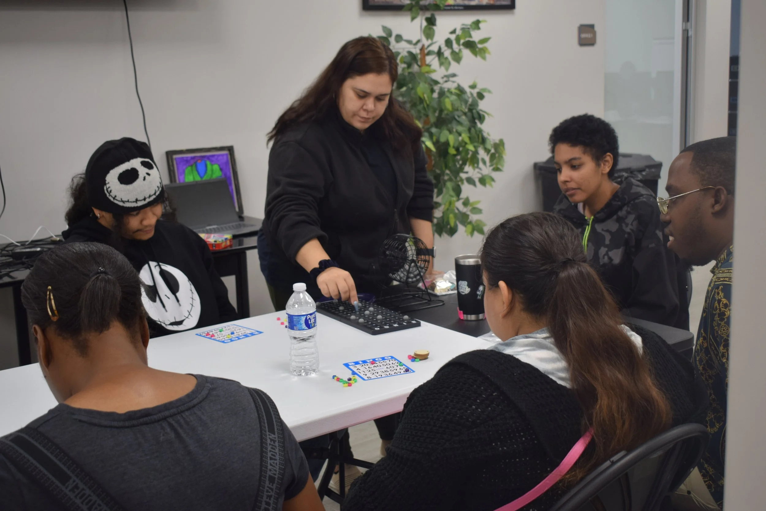 A group of six diverse young people playing a board game at a white table in a room, with a woman standing and controlling the game, and a laptop and drawing tablet on a desk in the background.