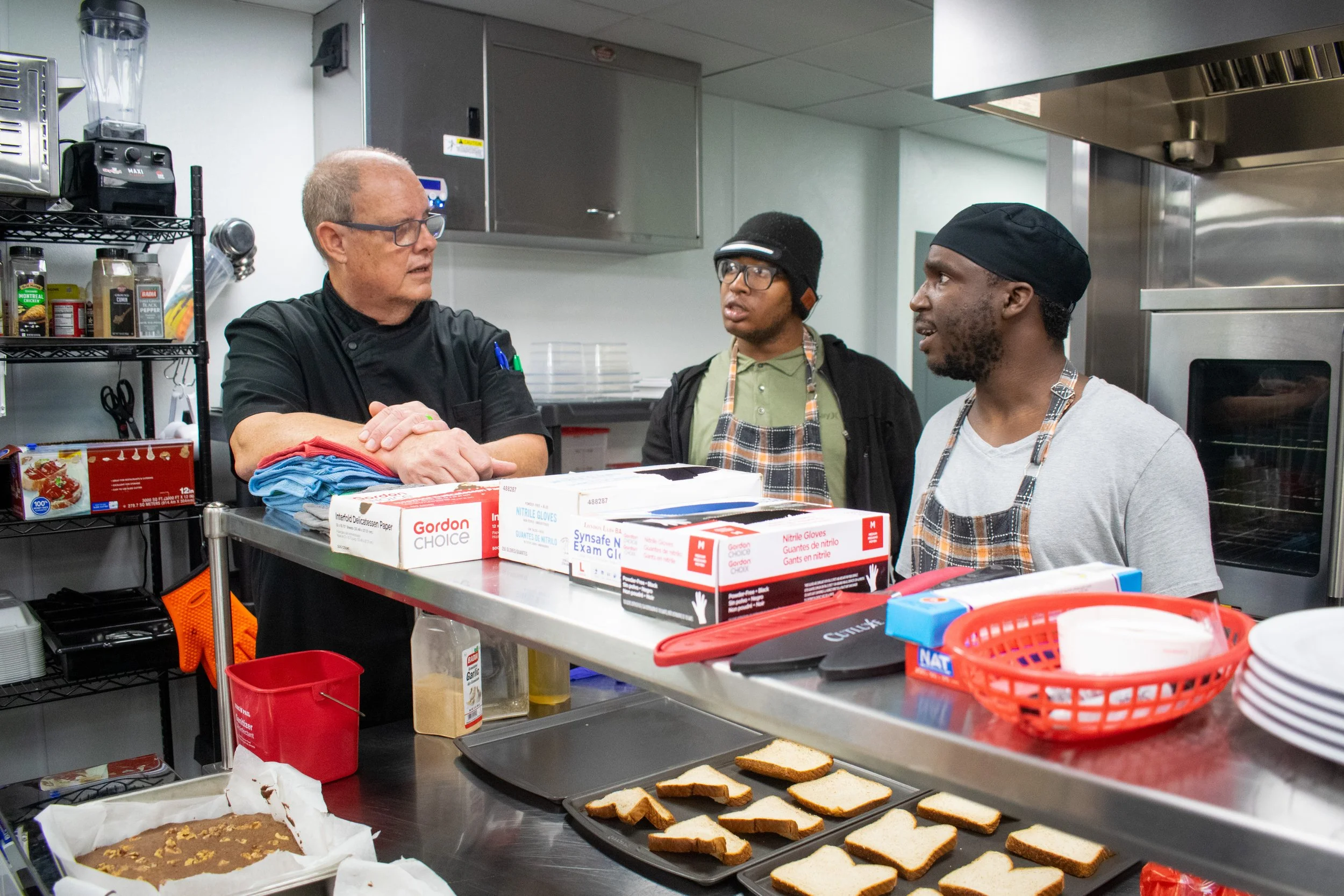 Three men in a commercial kitchen engaged in conversation. One is wearing glasses and a black chef coat, the other two are wearing aprons and hats. The counter has slices of bread and a partially baked loaf, with various baking supplies and tools around.