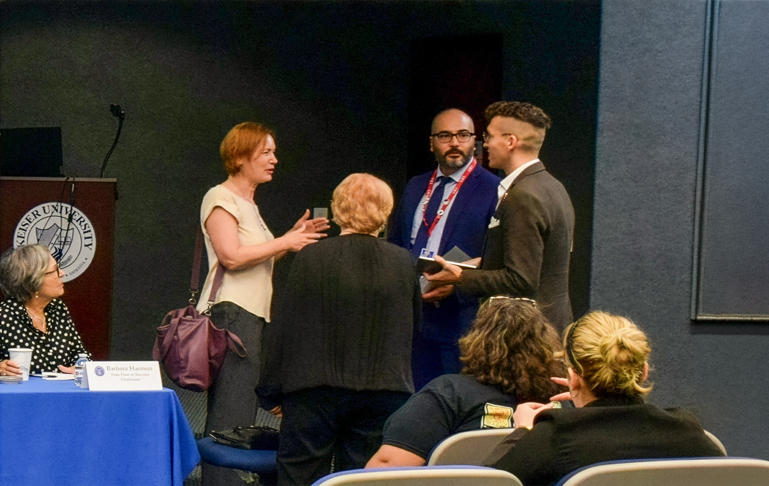 Group of five people engaged in conversation at a conference, with a podium and university logo in the background.