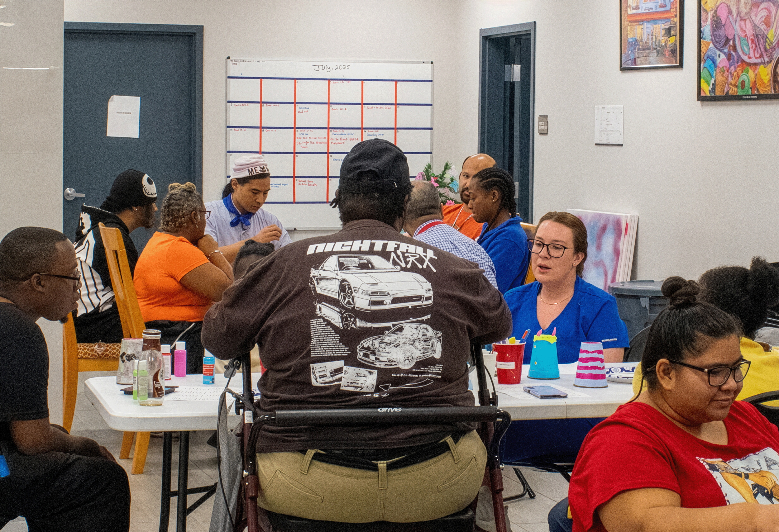 A group of diverse people sitting at a table in a room, engaged in a discussion or activity. The table has colorful cups, a smartphone, and various small items. There is a whiteboard with a calendar or schedule behind them, and framed artwork on the walls.