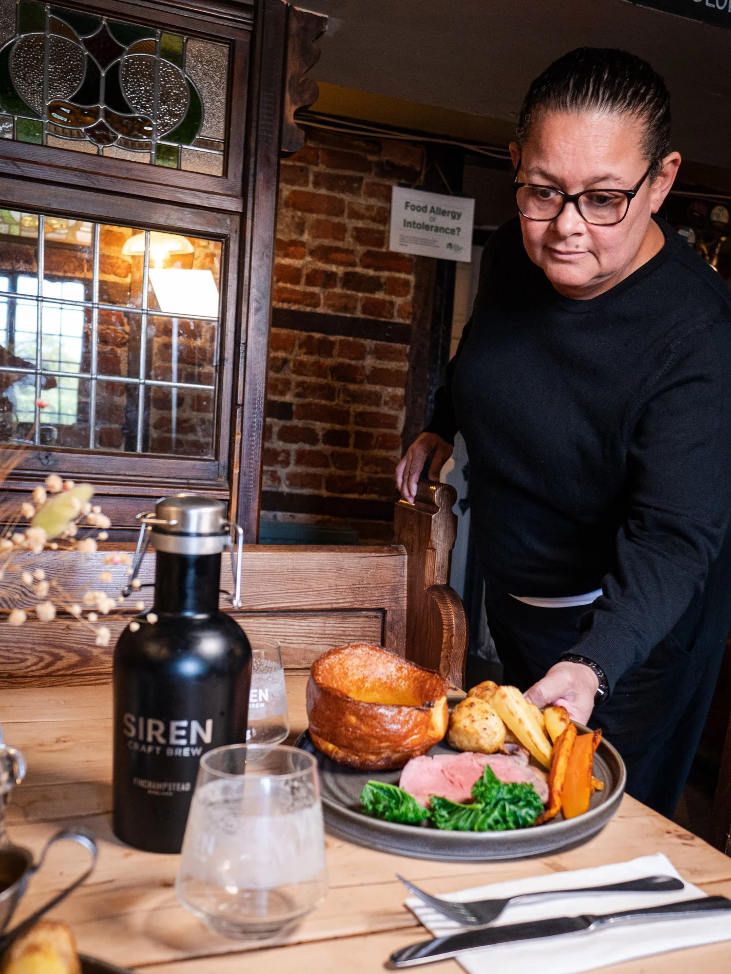 A person serving a plate of food in a restaurant with brick walls, a wooden table, and a bottle labeled 'Siren Craft Brew'. The plate contains baked ham, cooked carrots, potatoes, and greens.