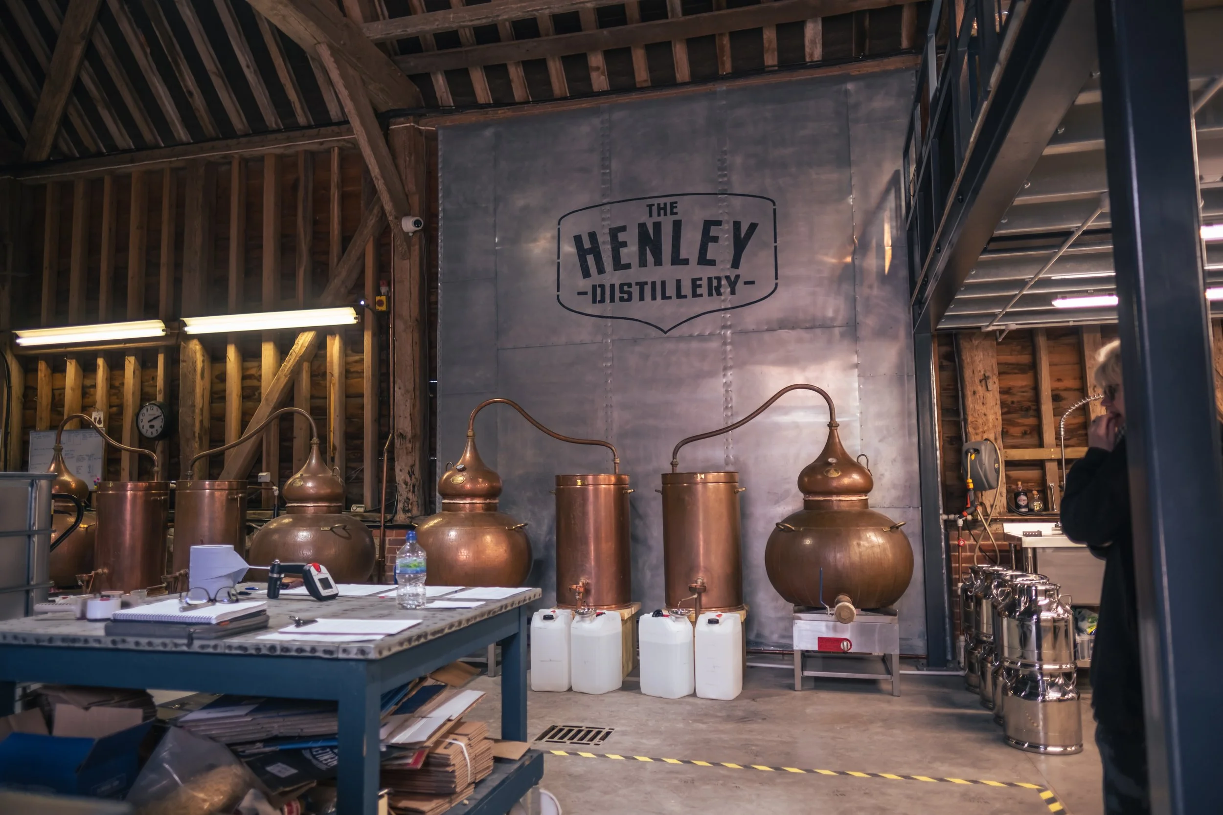 Inside The Henley Distillery with copper distillation apparatus, a work table with notes and a water bottle, and a person on the right talking on the phone.