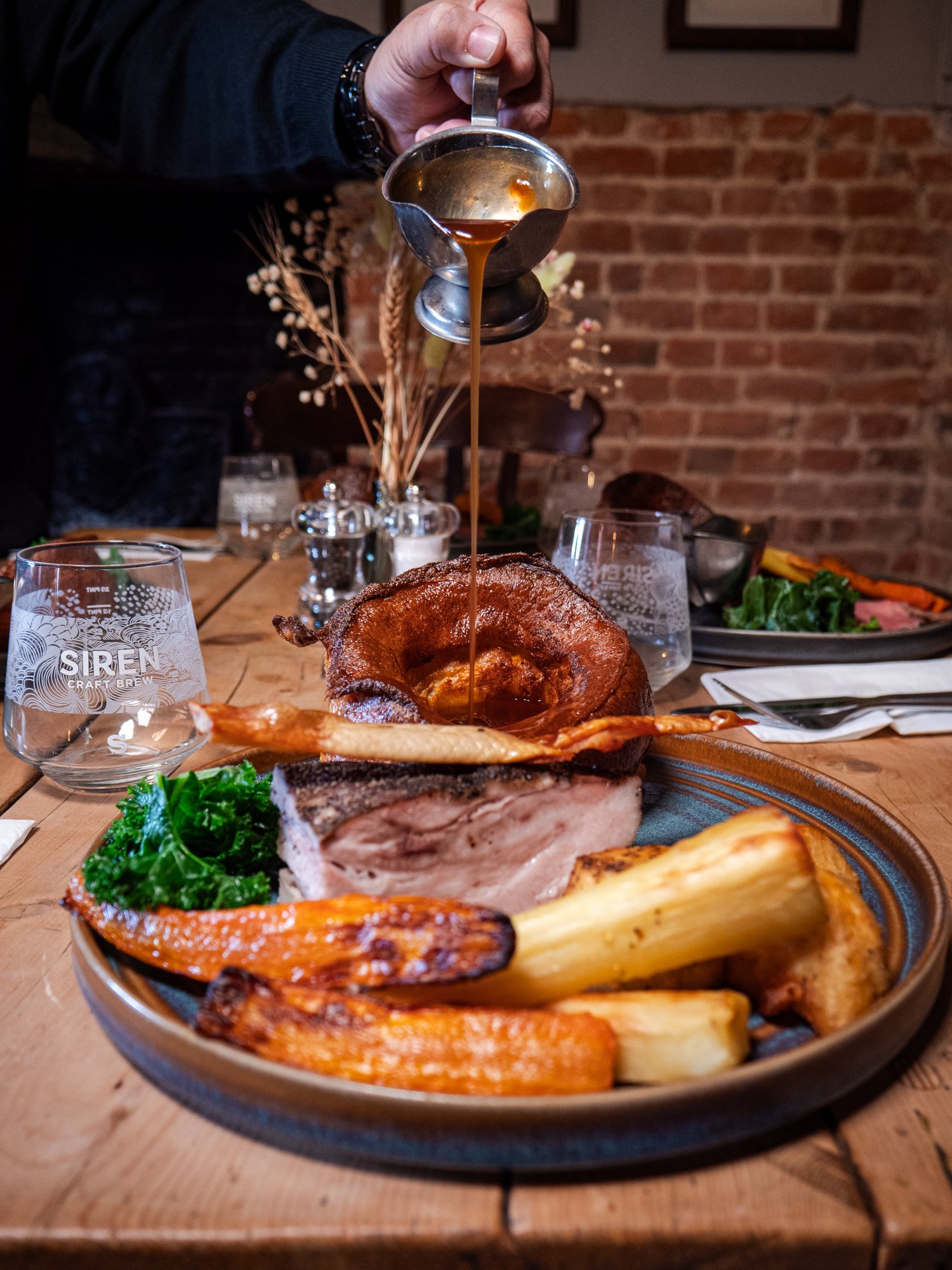 Person pouring gravy over a plate of roasted meat, vegetables, and greens at a restaurant.