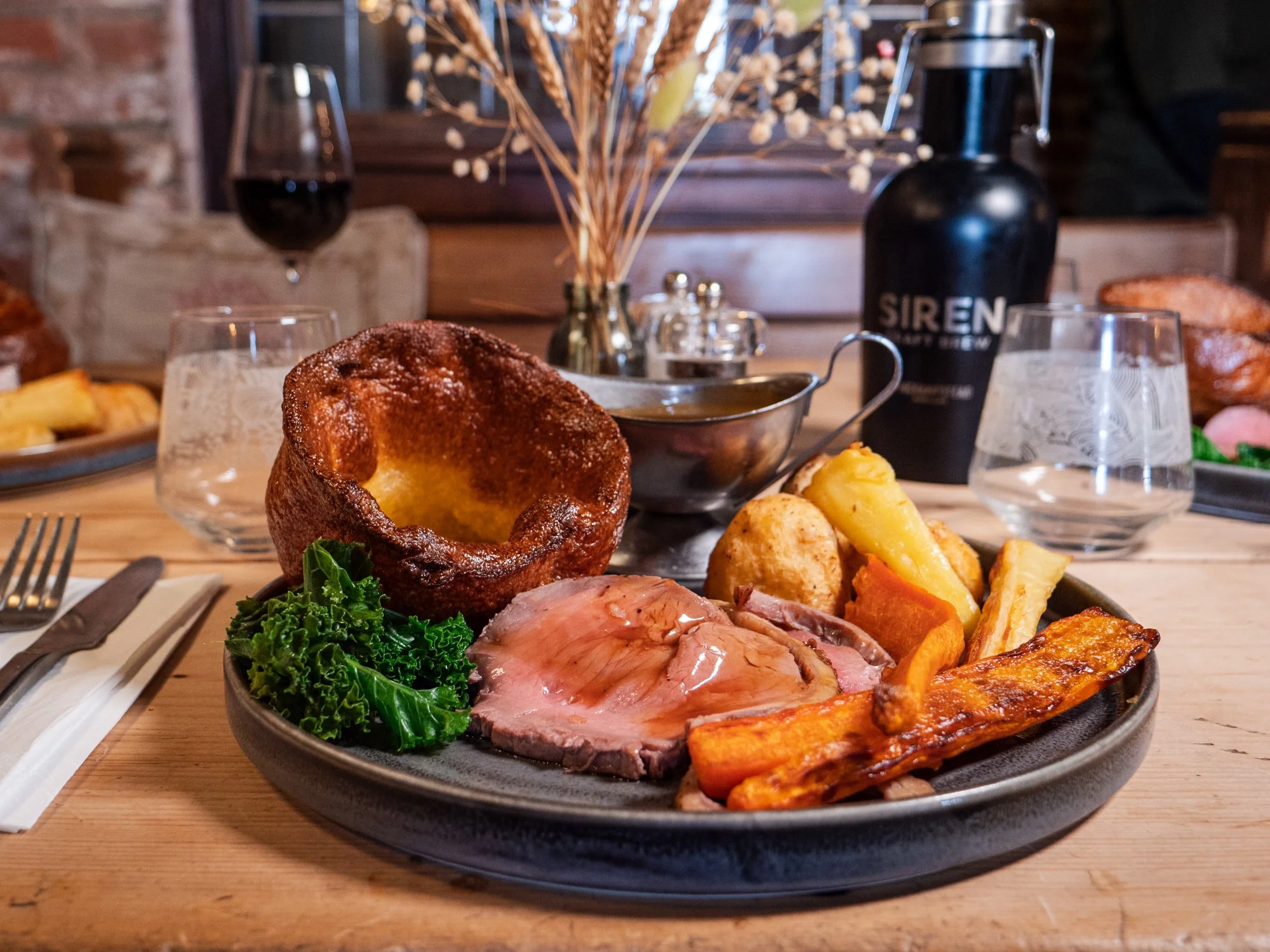 A plate of roast beef, roast potatoes, roasted root vegetables, kale, Yorkshire pudding, and a pork rib, with a glass of red wine and a glass of water on a wooden table in a rustic restaurant setting.