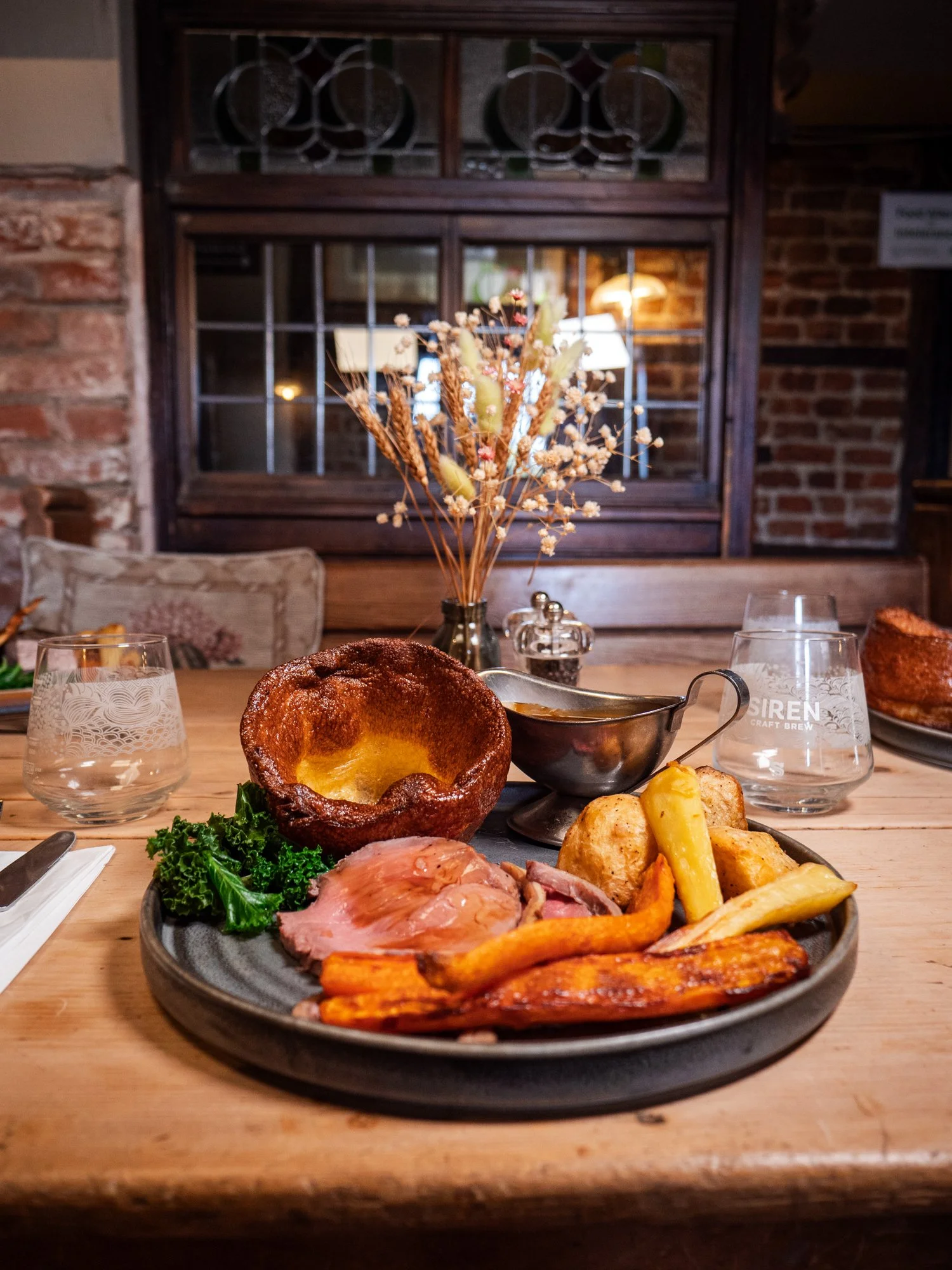 Plate of roast beef with vegetables, Yorkshire pudding, and potatoes on a wooden table, with glass water glasses and a decorative vase in the background.