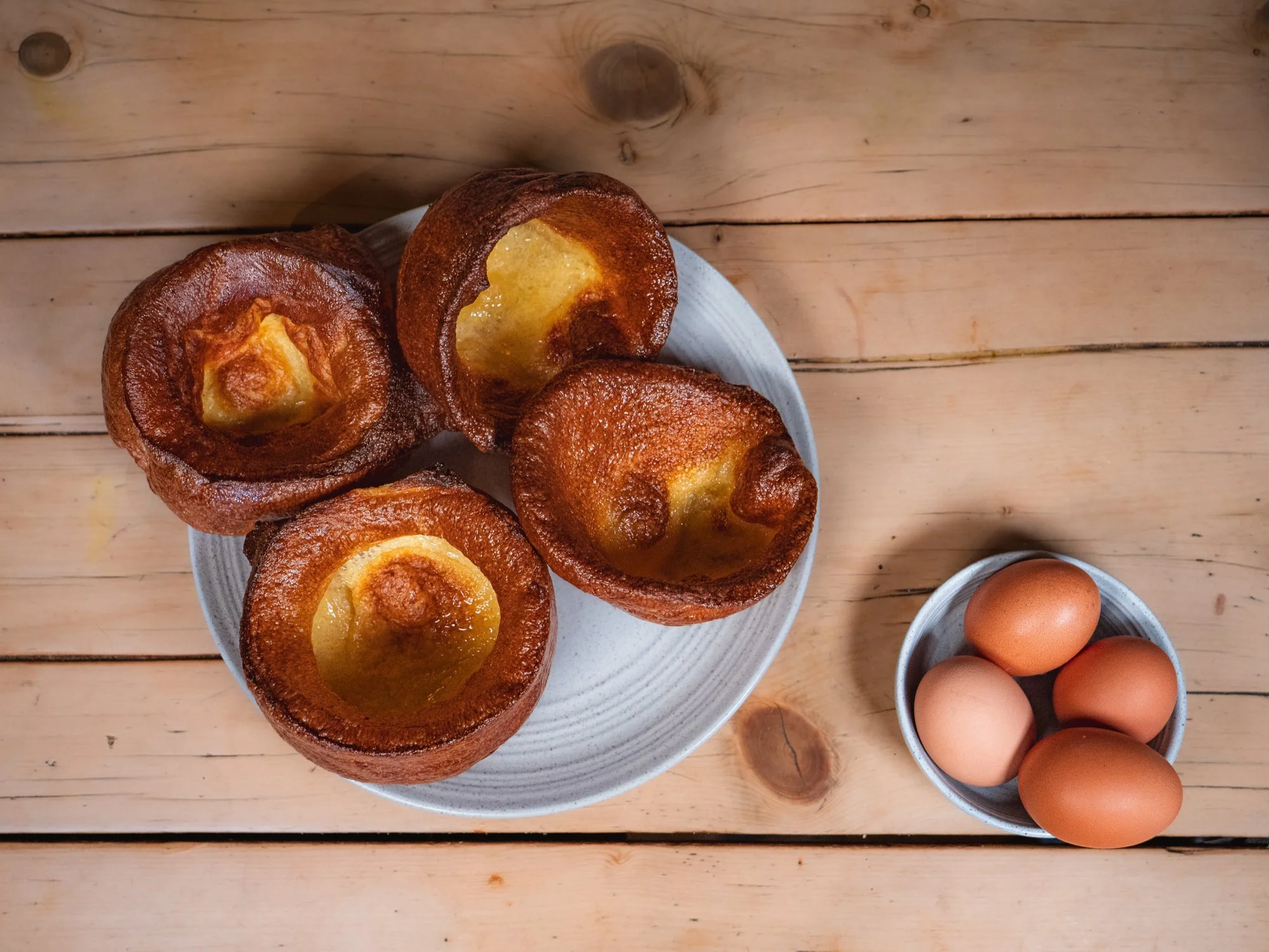 Four baked egg-centered pastries on a white plate with a small bowl of brown eggs beside it on a wooden surface.