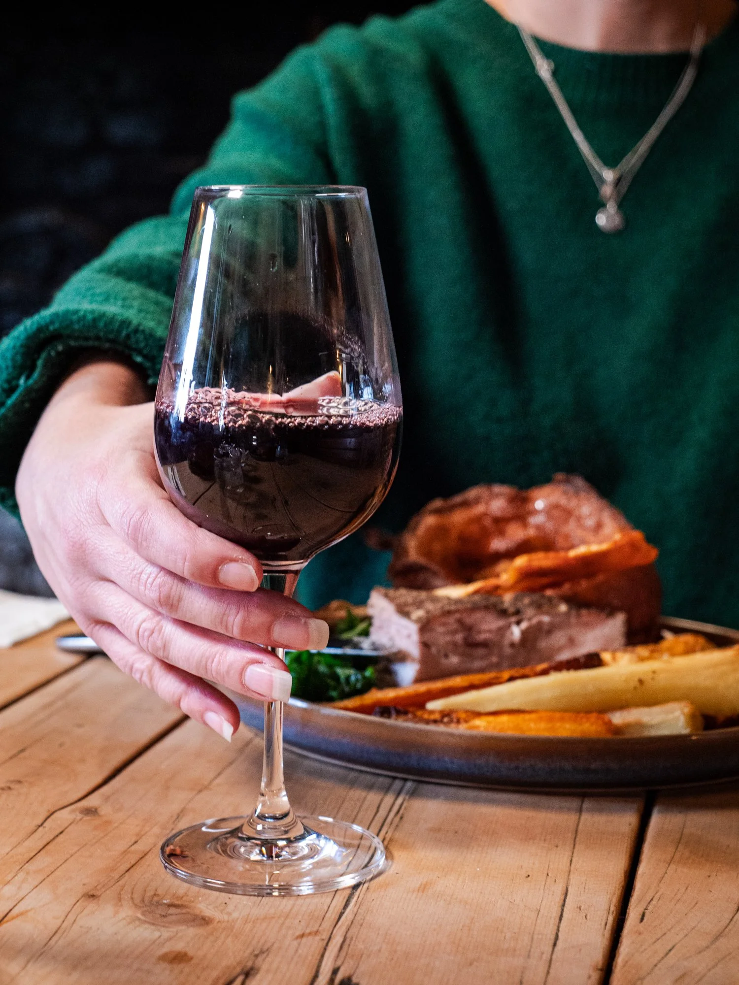 Person holding a glass of red wine with a plate of food, including meat and vegetables, on a wooden table.