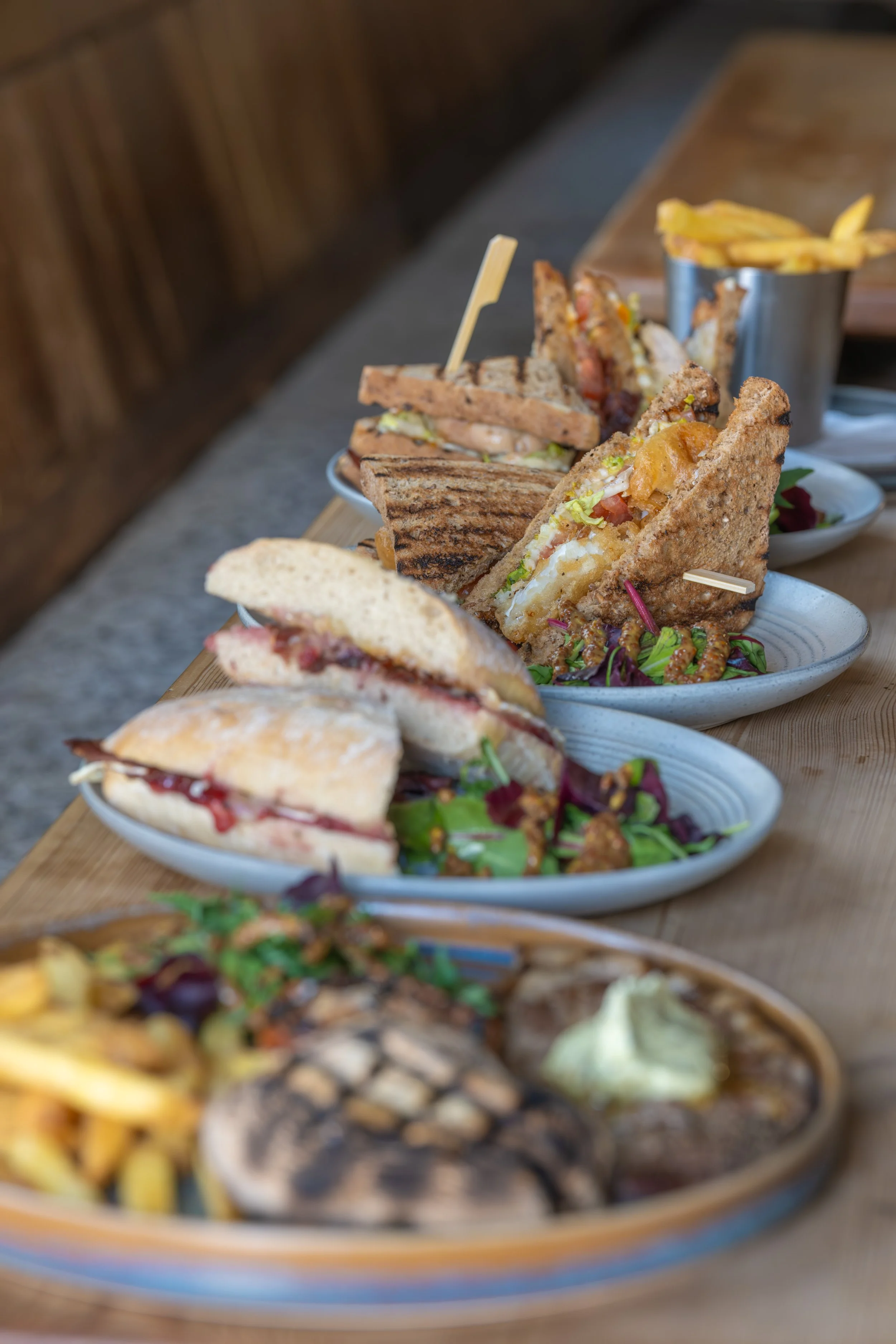 Assorted sandwiches, fries, and snacks displayed on plates on a wooden table at a restaurant.