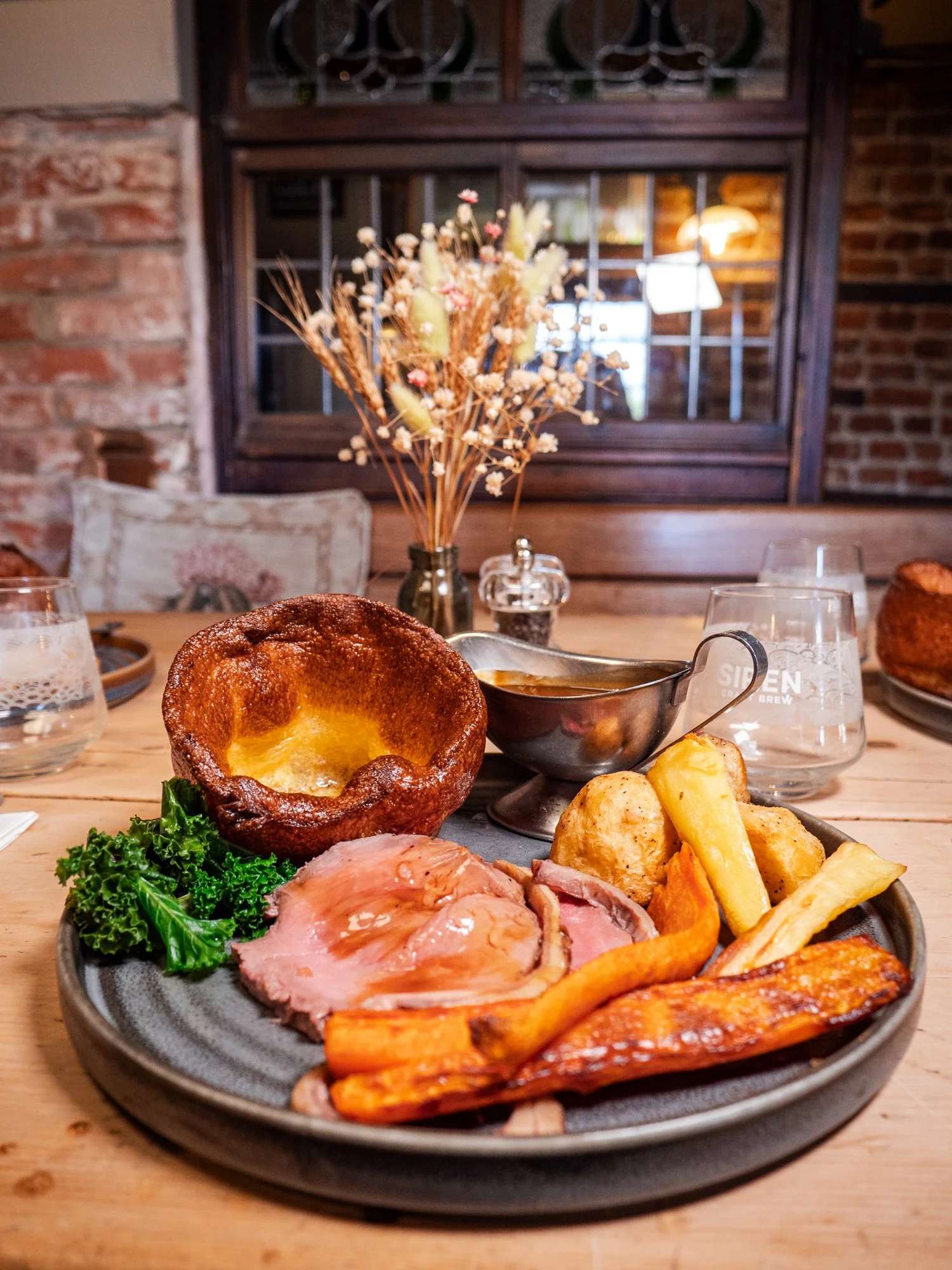 A plate of roast beef, Yorkshire pudding, roasted vegetables, and greens on a wooden table in a cozy restaurant setting.