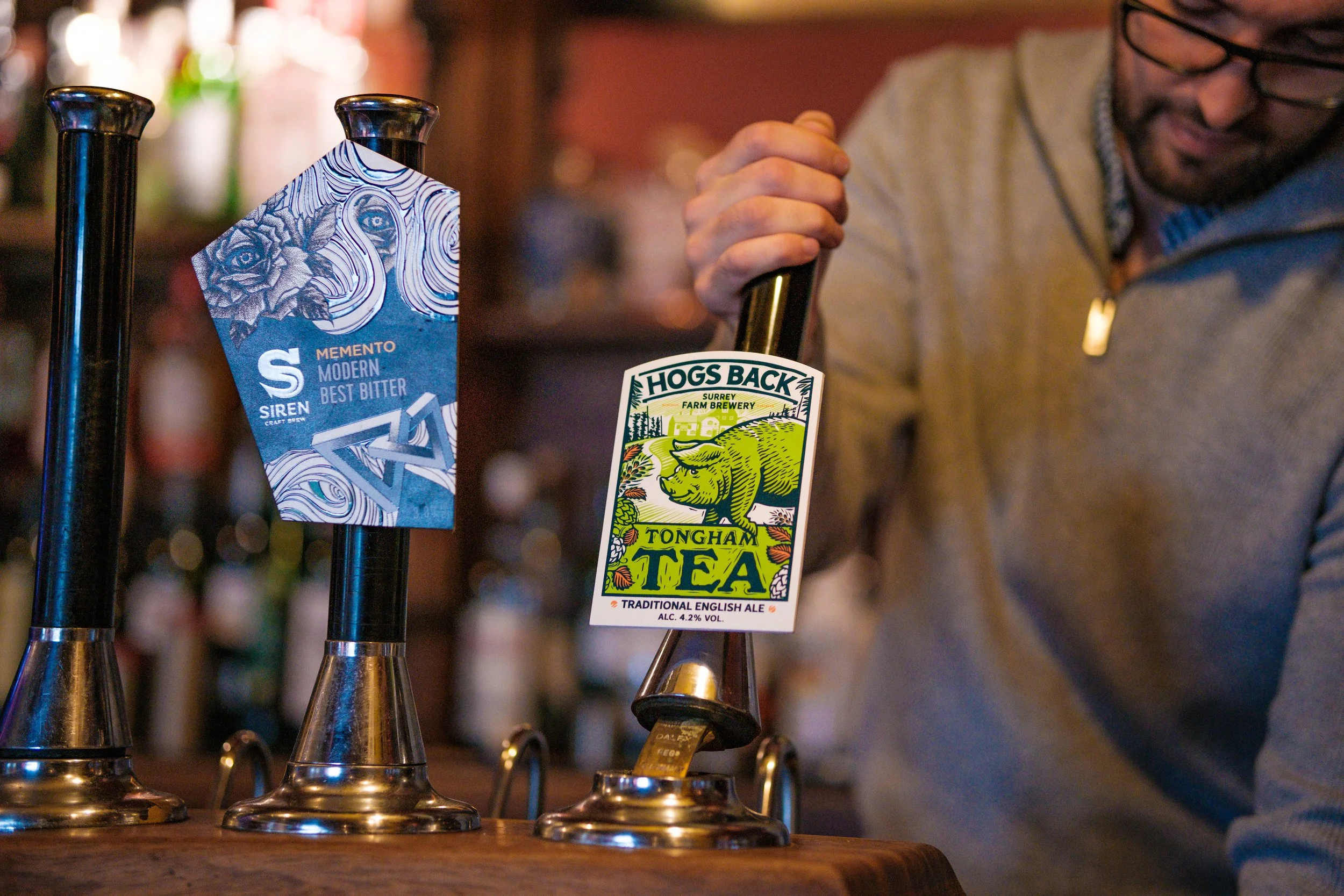A person pouring craft beer from a tap labeled Hog's Back Tongham Tea at a bar. There are two taps, one labeled Siren Memento Modern Best Bitter and the other Hog's Back Tongham Tea.