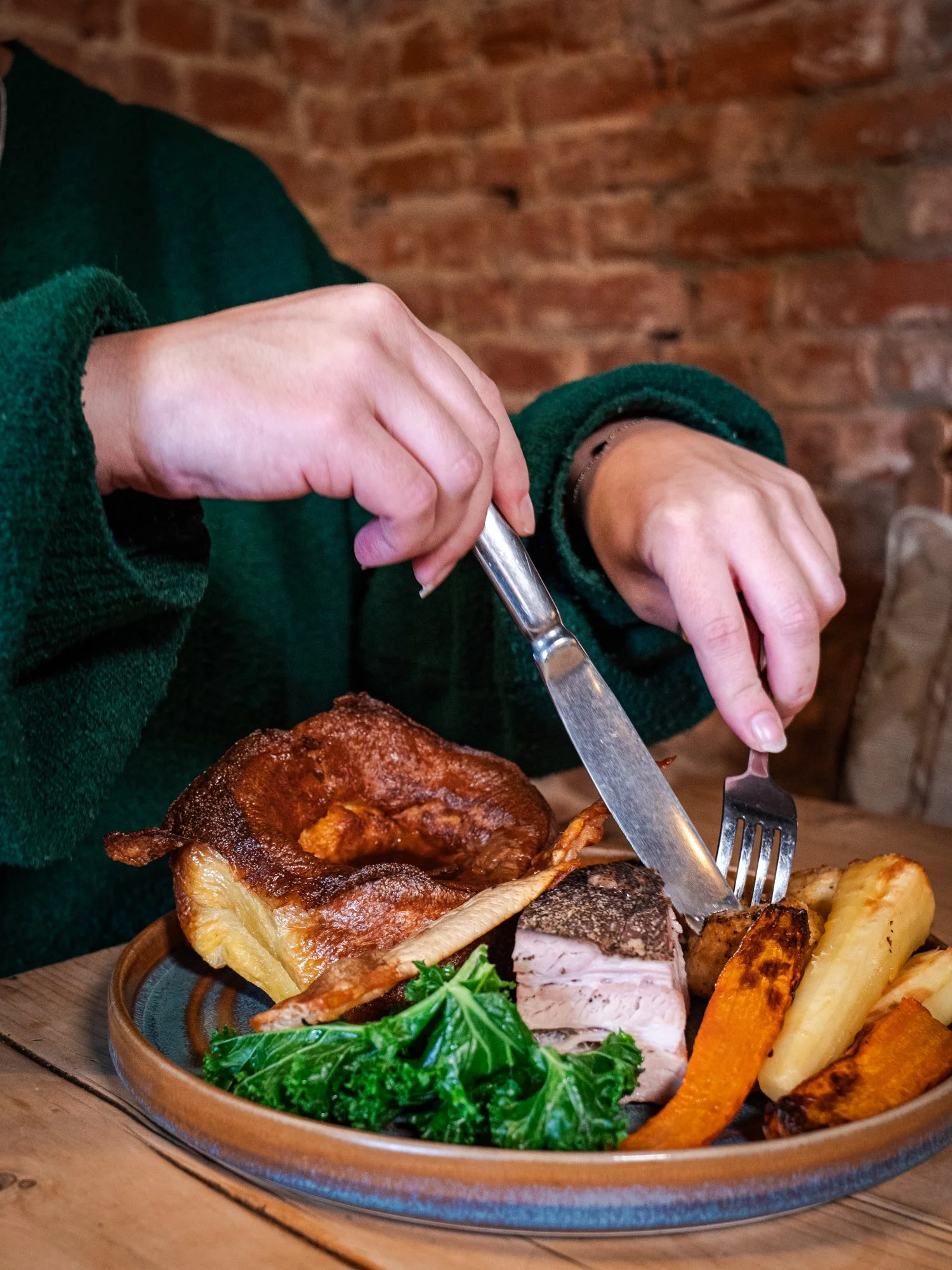 Person in a green sweater cutting a plated meal with roasted meat, vegetables, and greens, set against a brick wall background.