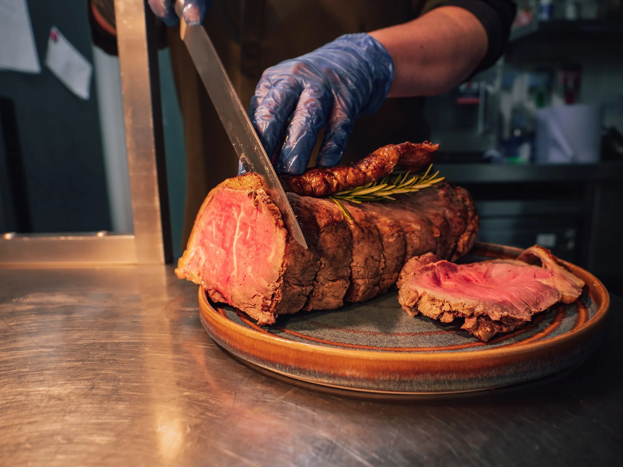 Person slicing a cooked large roast beef with a large knife, wearing blue plastic gloves, on a decorative plate with a sprig of rosemary on top.