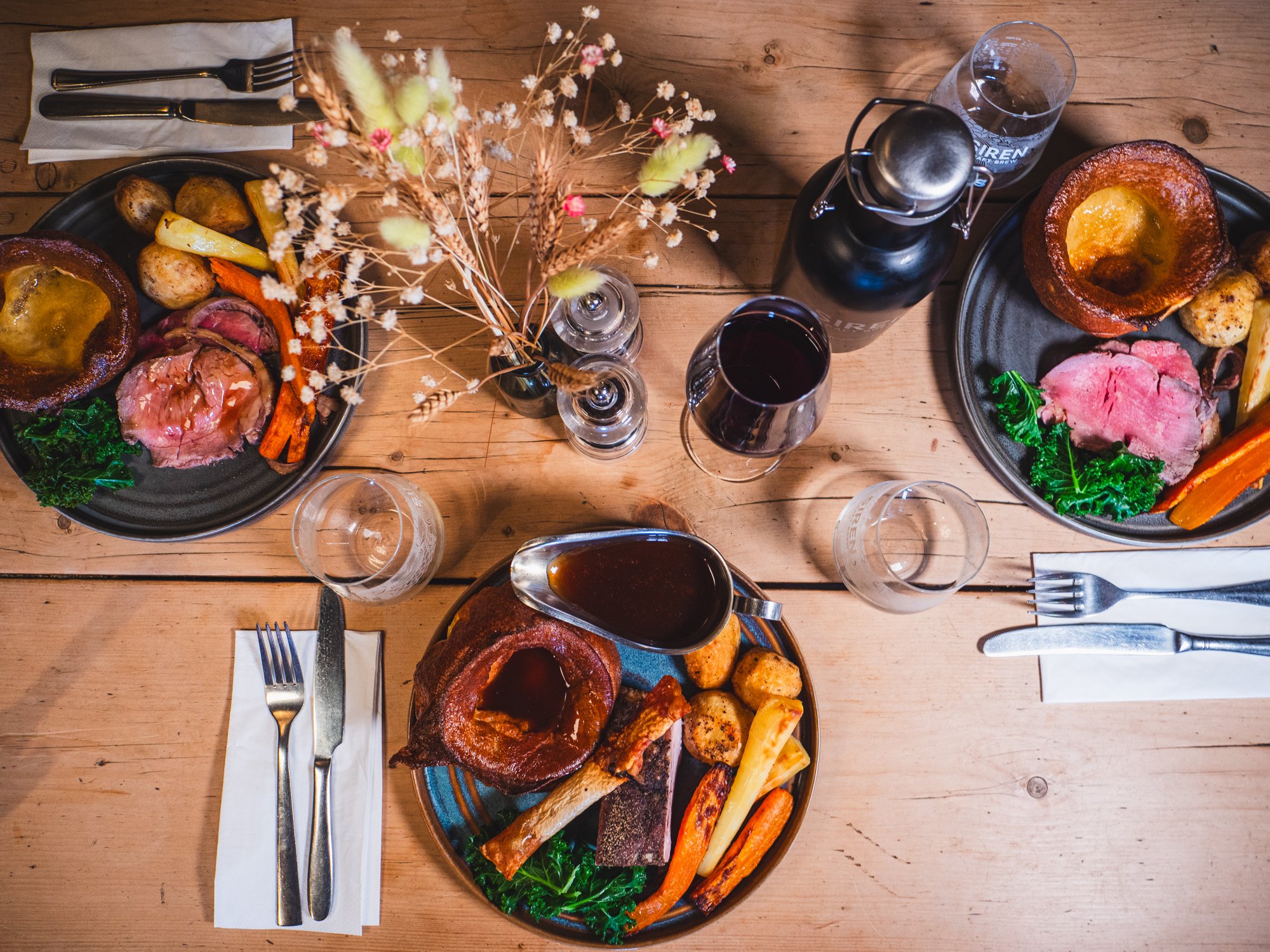 Three plates of a roast dinner with roast beef, Yorkshire pudding, roasted vegetables and potatoes on a wooden table, with wine, water, and a flower arrangement in the center.