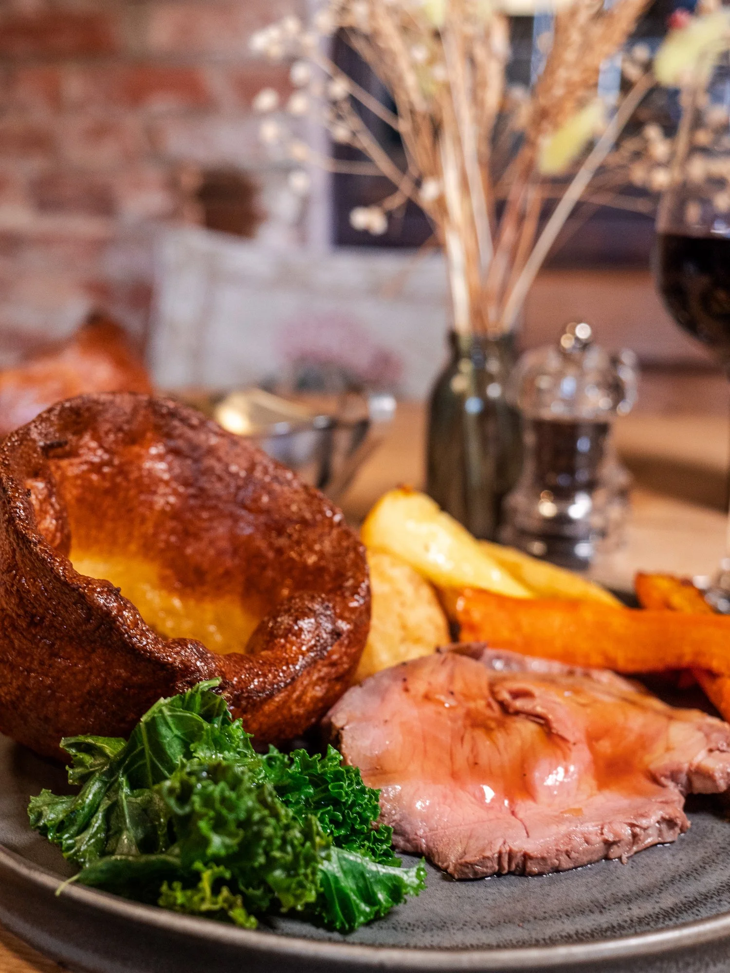Plate of food with Yorkshire pudding, roast beef slices, roasted potatoes, carrots, and greens with a glass of red wine in the background.