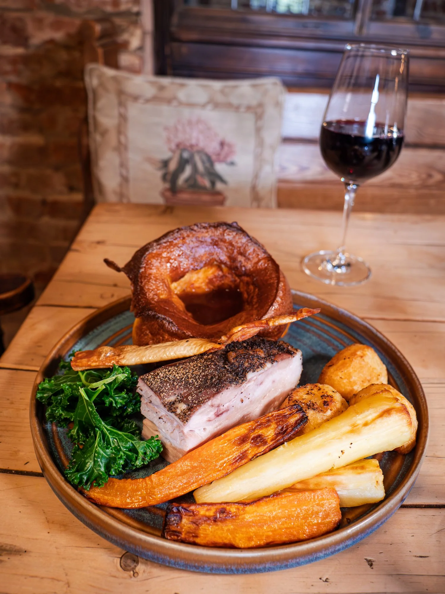 A rustic plate of food including baked fish, roasted root vegetables, kale, a slice of bacon, fried onion, and a large onion ring, with a glass of red wine on a wooden table in a cozy restaurant setting.