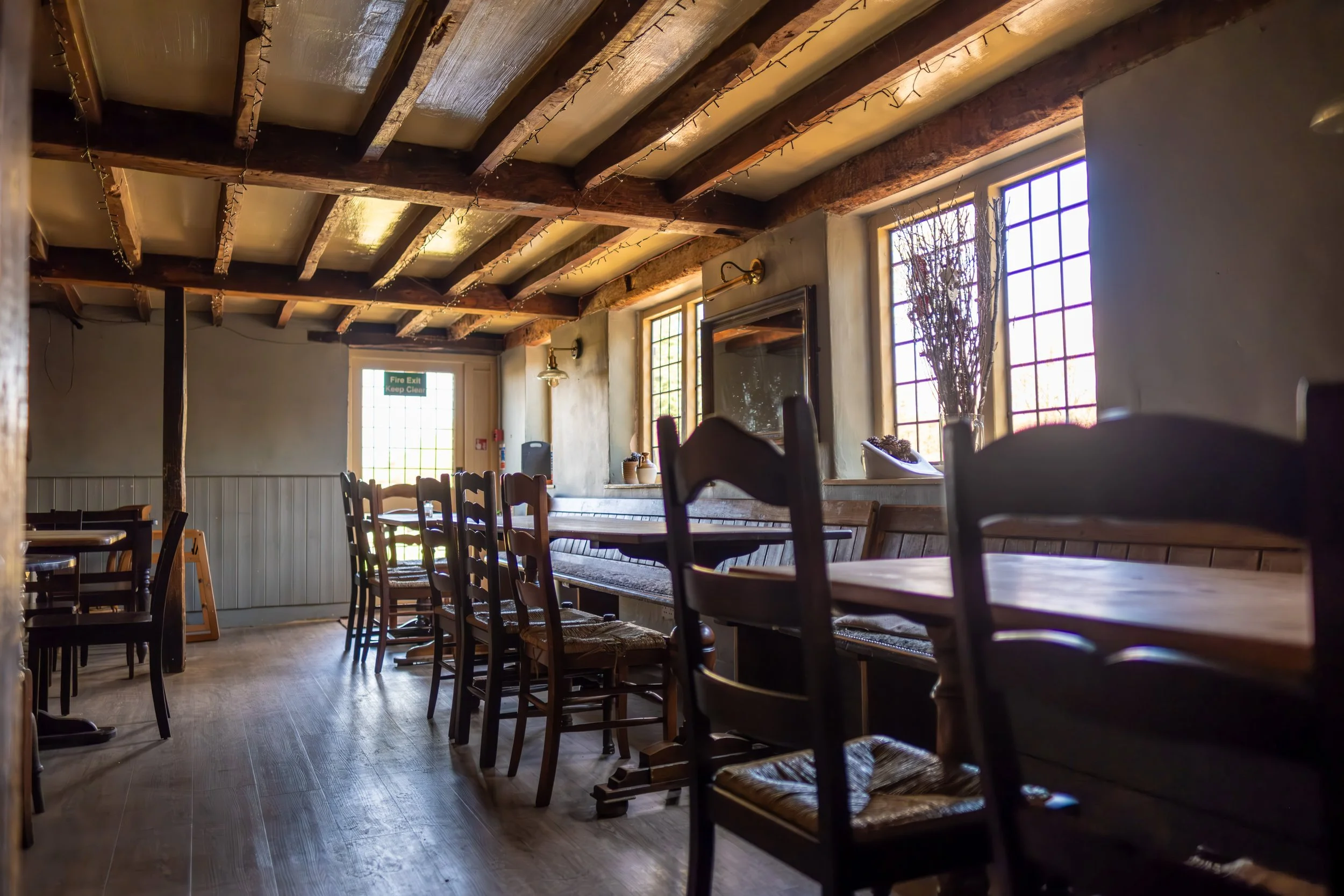 Interior of a cozy, rustic restaurant with wooden tables and chairs, natural light coming through large grid-patterned windows, and decorative items such as a large vase with dried flowers.