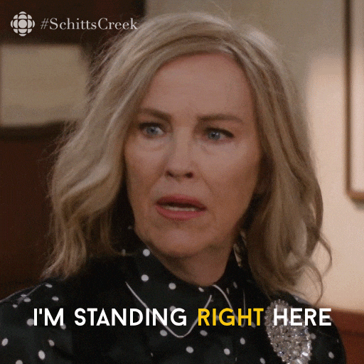 A woman with blonde hair wearing a black polka dot shirt and a brooch speaks in a scene from Schitt's Creek, with the text 'I'm standing right here.'