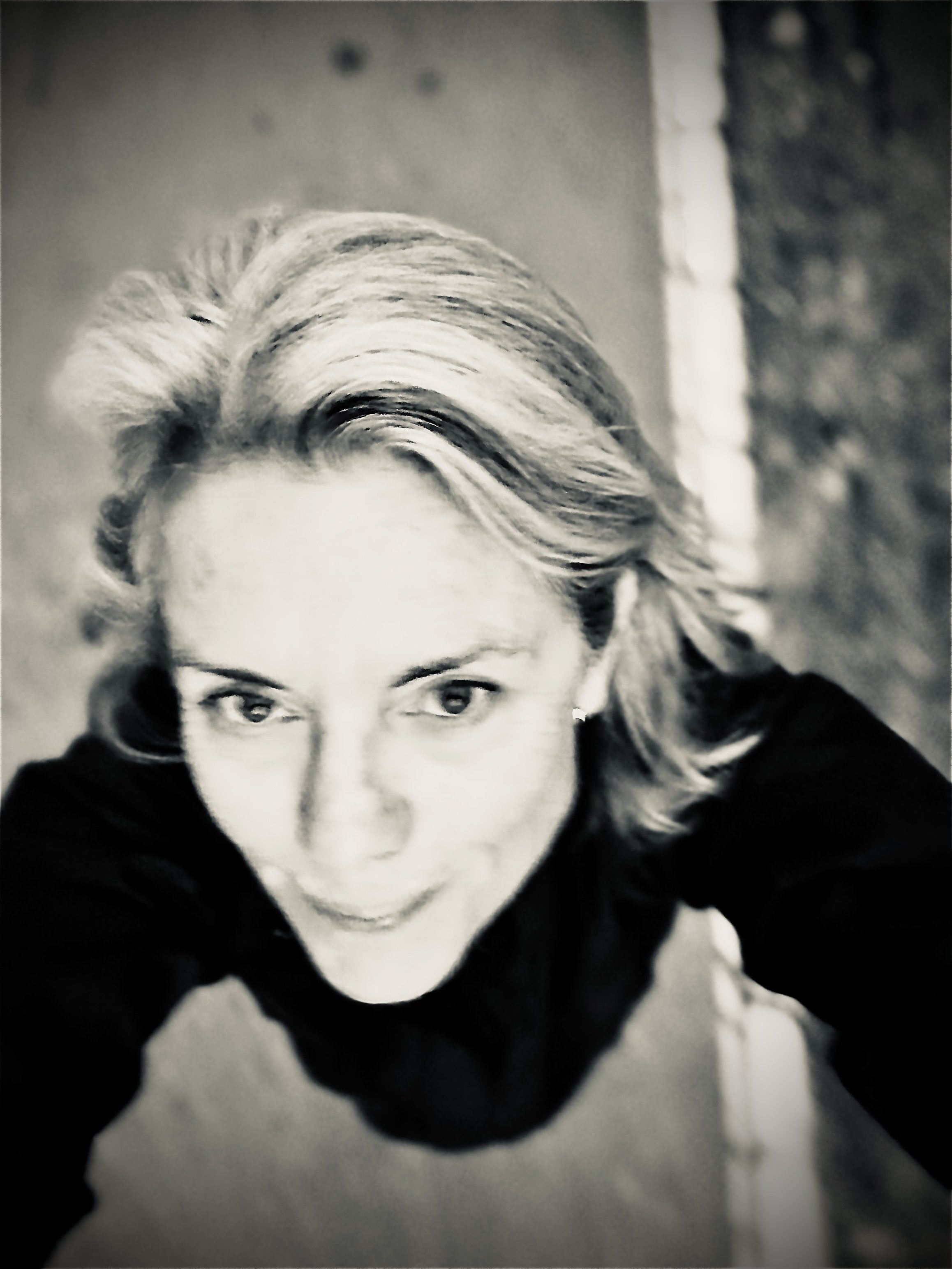 Black and white selfie of a woman with wavy blonde hair, smiling slightly, taken from an overhead angle.